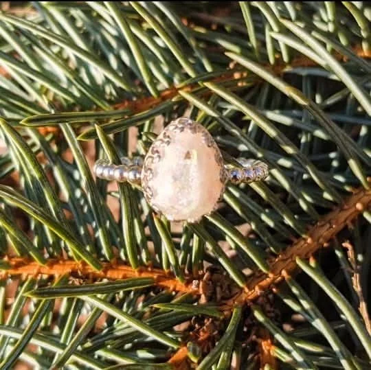 A silver ring with a large, oval-shaped pearl surrounded by small diamonds, resting on green pine needles.