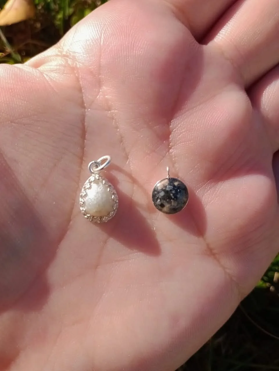 Hand holding two spherical stone pendants, one white and one black, with metal loops for jewelry attachment.