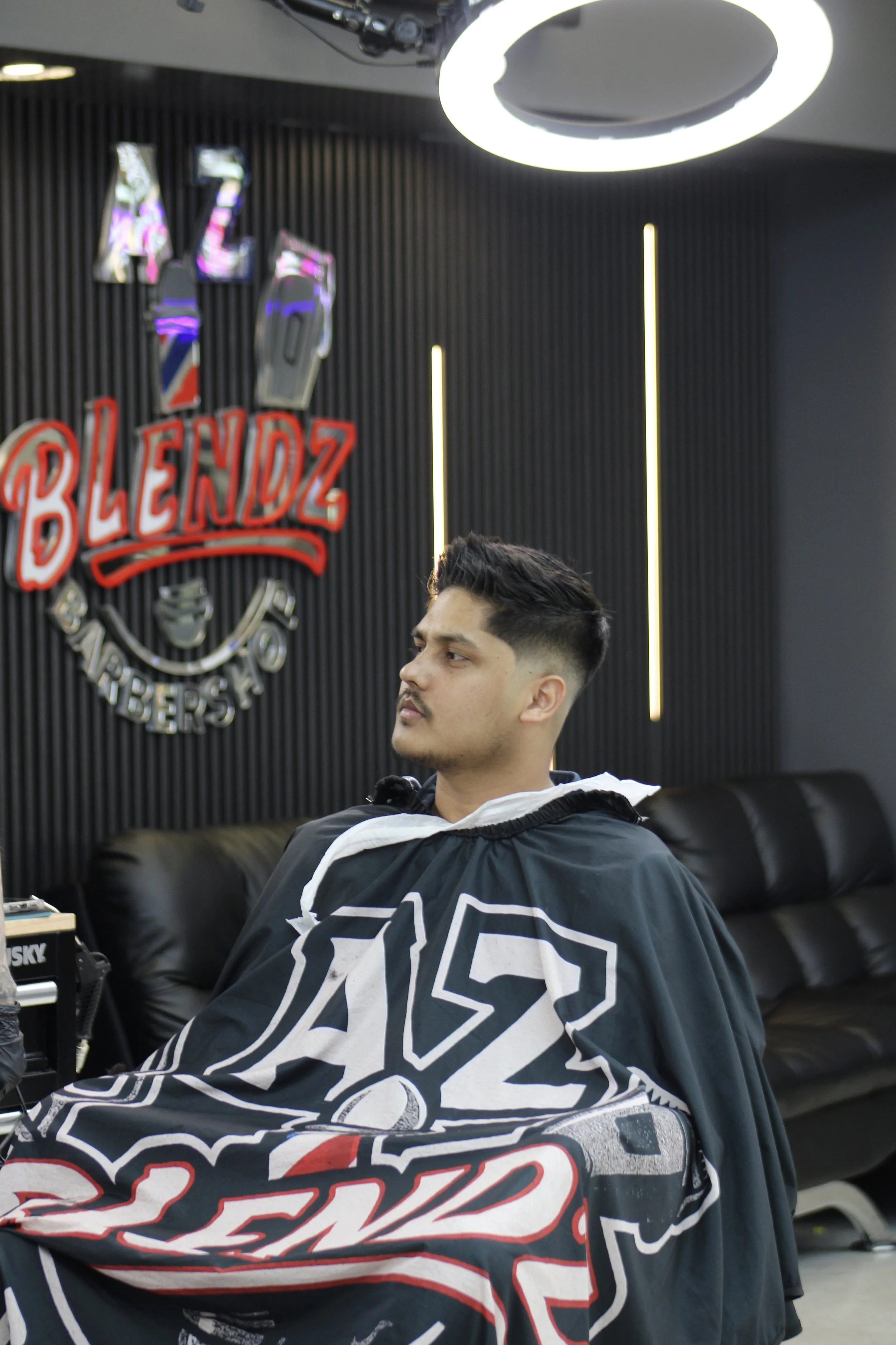 A man sitting in a barbershop chair with a black and white barber cape. The shop has a black wall with a circular ceiling light and a neon sign that says 'BLENDZ' with other barber-related images.