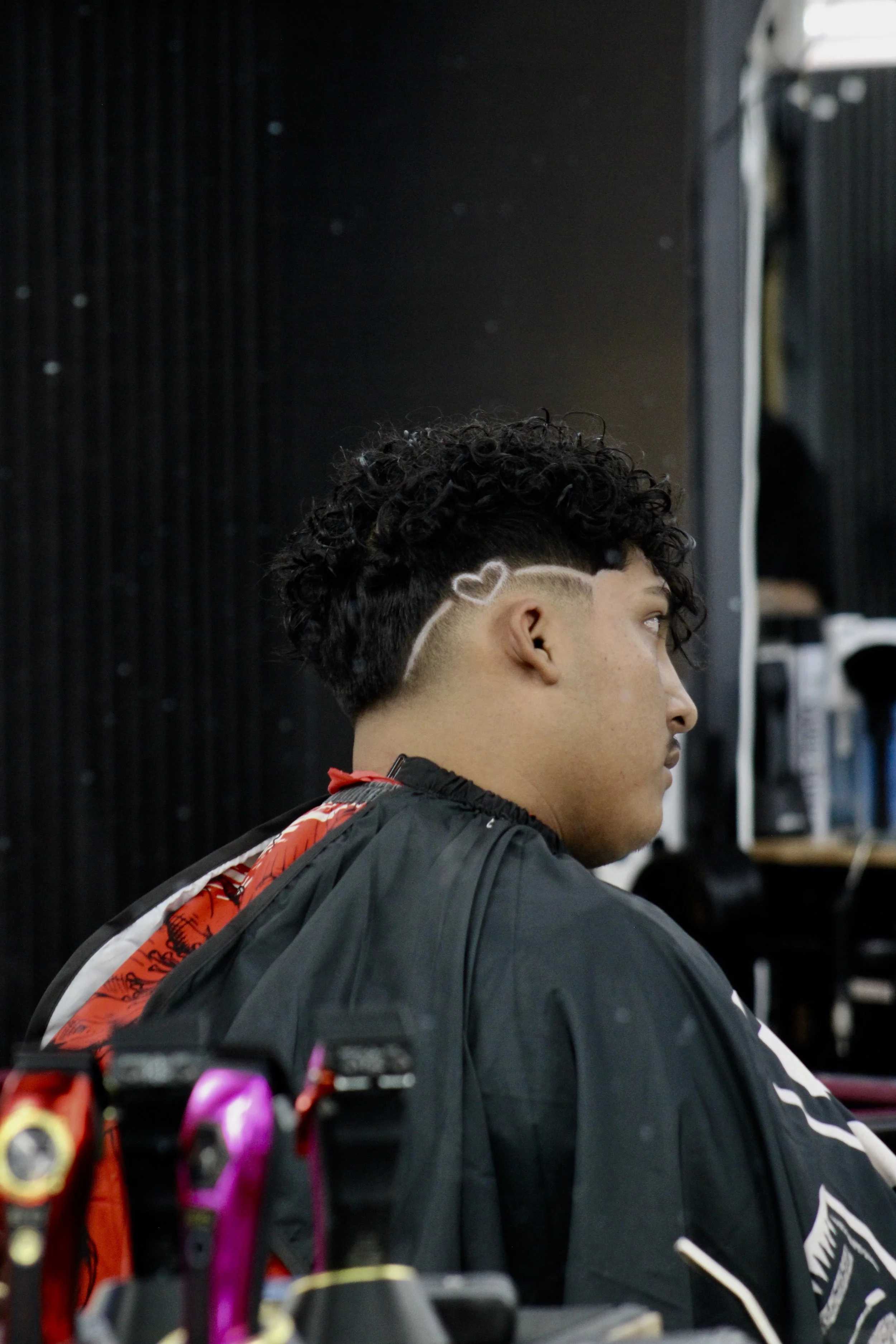 A young man getting a haircut at a barbershop, seated with a black barber cape over his shoulders, facing to the right. He has dark, curly hair with a design shaved into the side of his head.