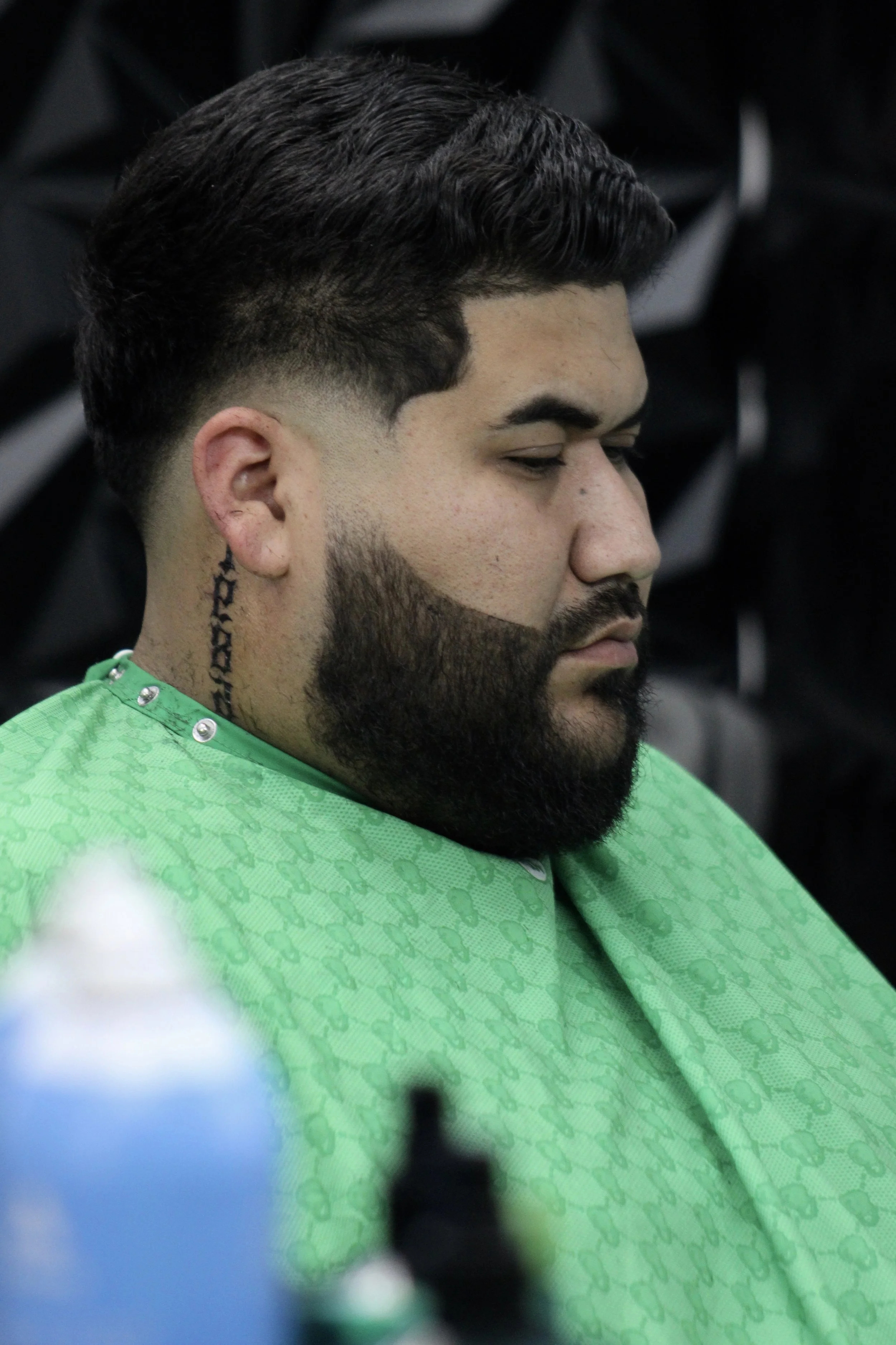 Profile of a man with dark, styled hair and a beard, sitting with eyes closed, wearing a green barber cape.