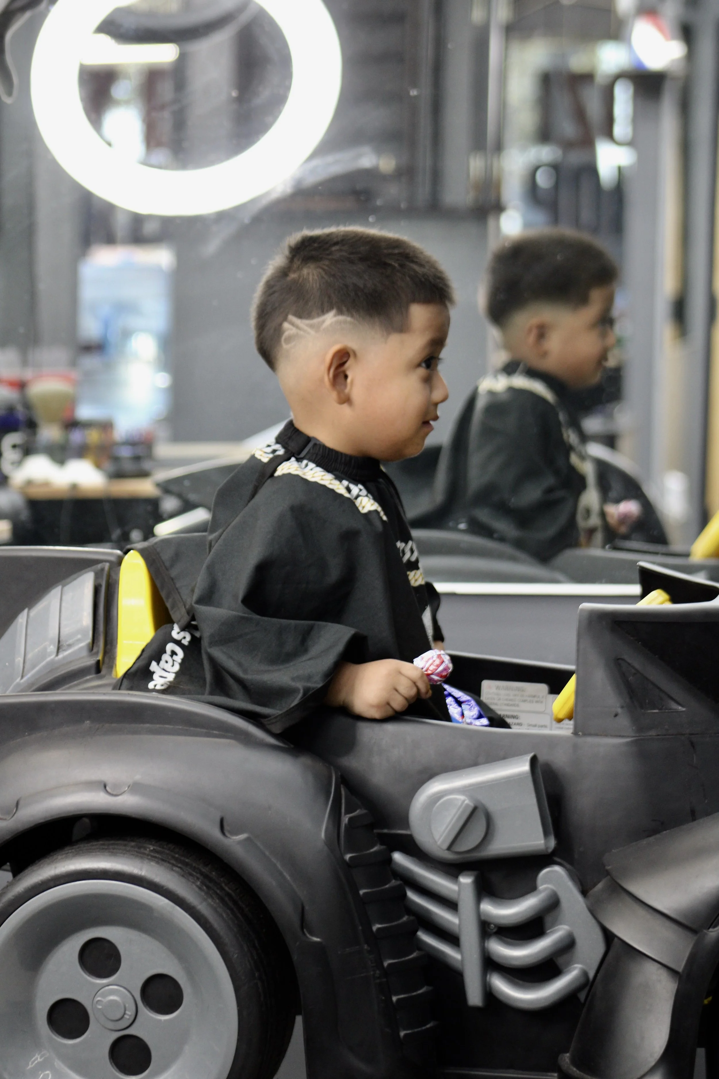 A young boy sitting in a black toy car that resembles a real vehicle, with a reflection of himself in the mirror behind him. The boy is holding a lollipop and wearing a black cape or jacket.
