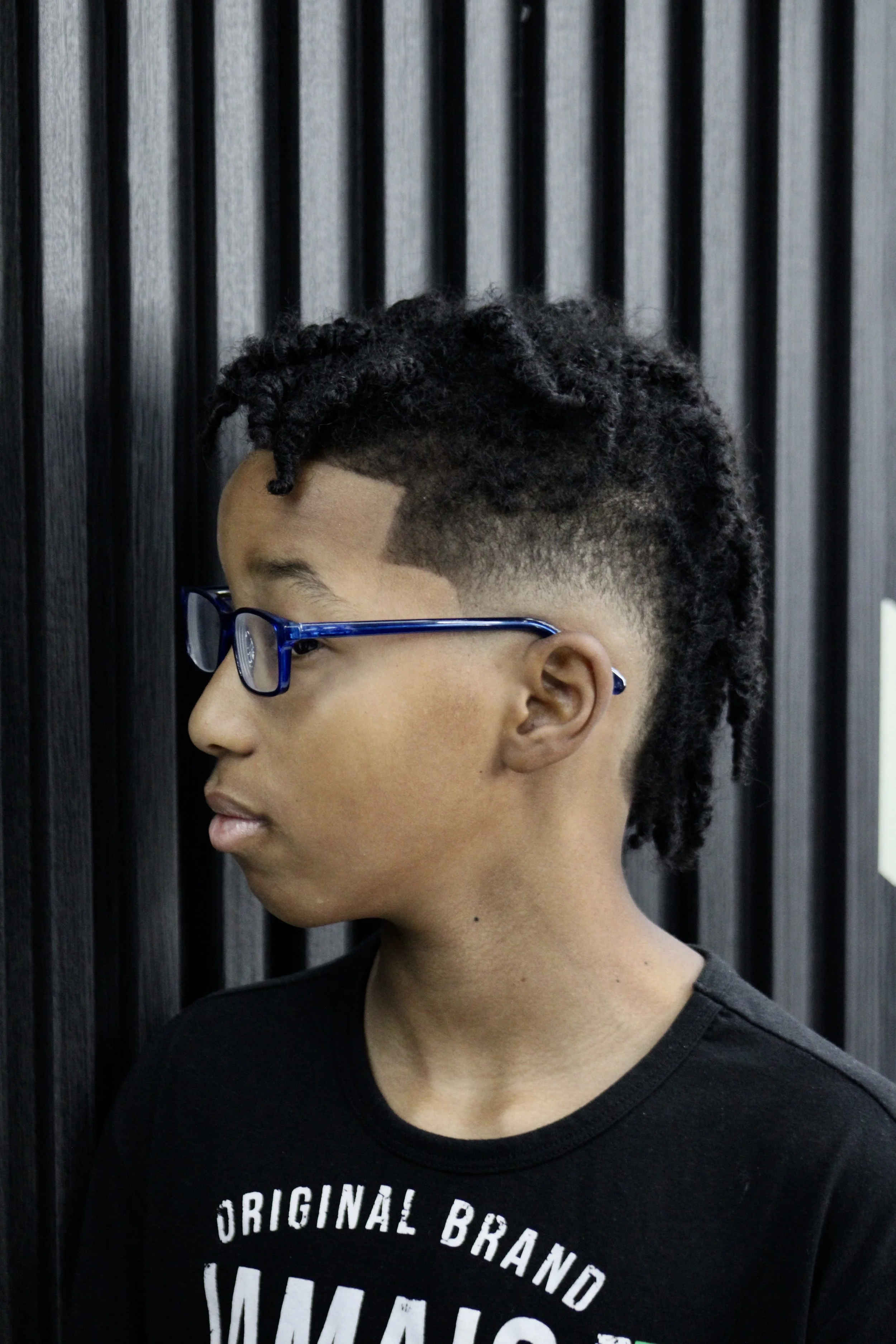 Side profile of a young person with styled dreadlocks and wearing blue glasses, standing against a black slatted wall.