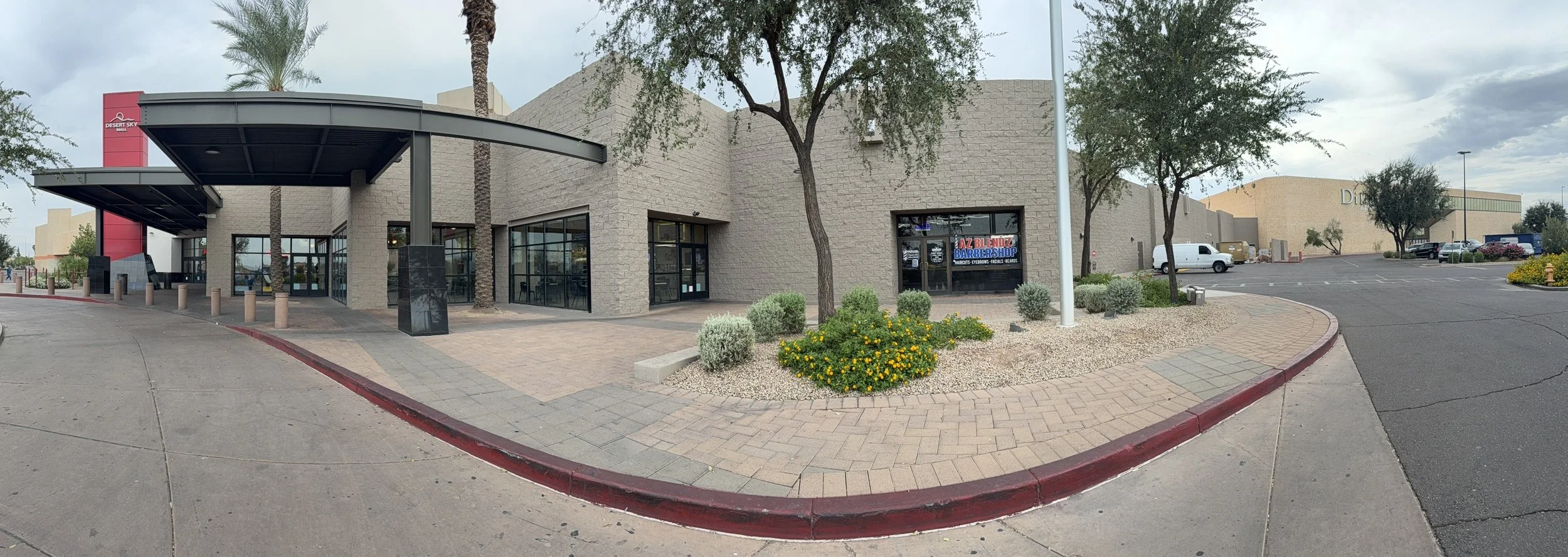 Panoramic view of a shopping mall exterior with a palm tree, flower bed, and storefronts, including a barbershop sign, under cloudy skies.
