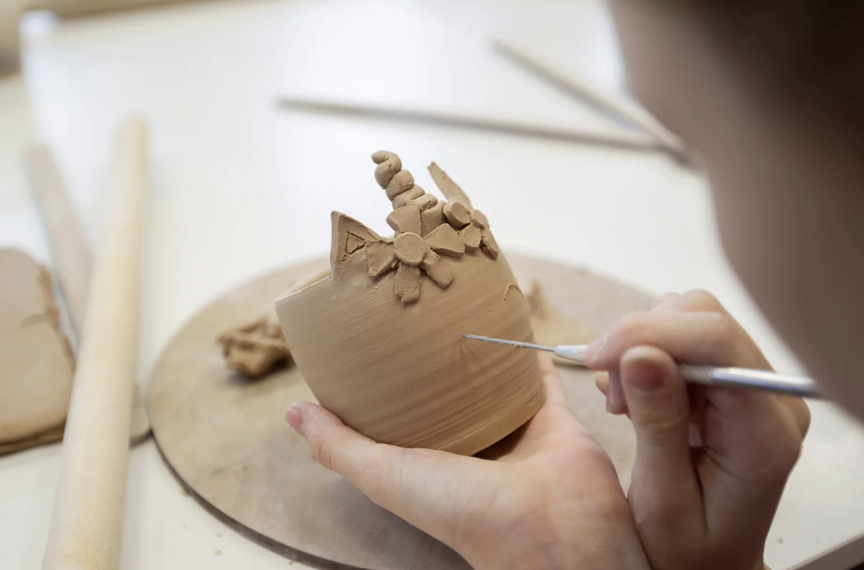 Person sculpting a clay unicorn head with a horn and ears, working on a pottery wheel, with sculpting tools and clay pieces visible.