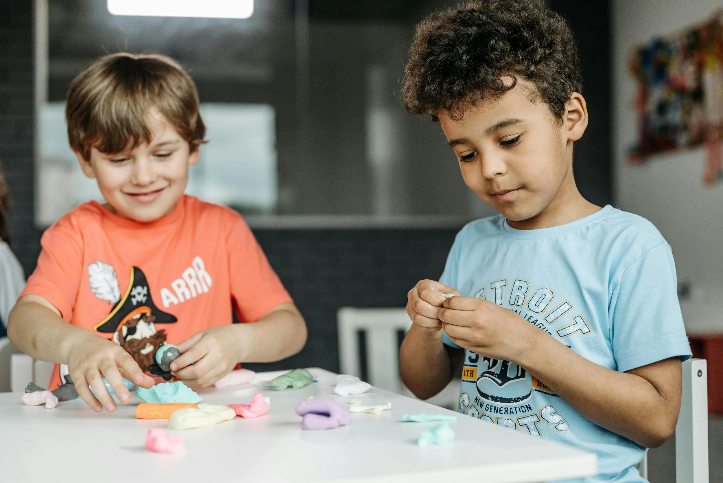 Two young children working with clay at a table, demonstrating hands-on sensory play, fine motor engagement, and emotional regulation in a classroom setting.