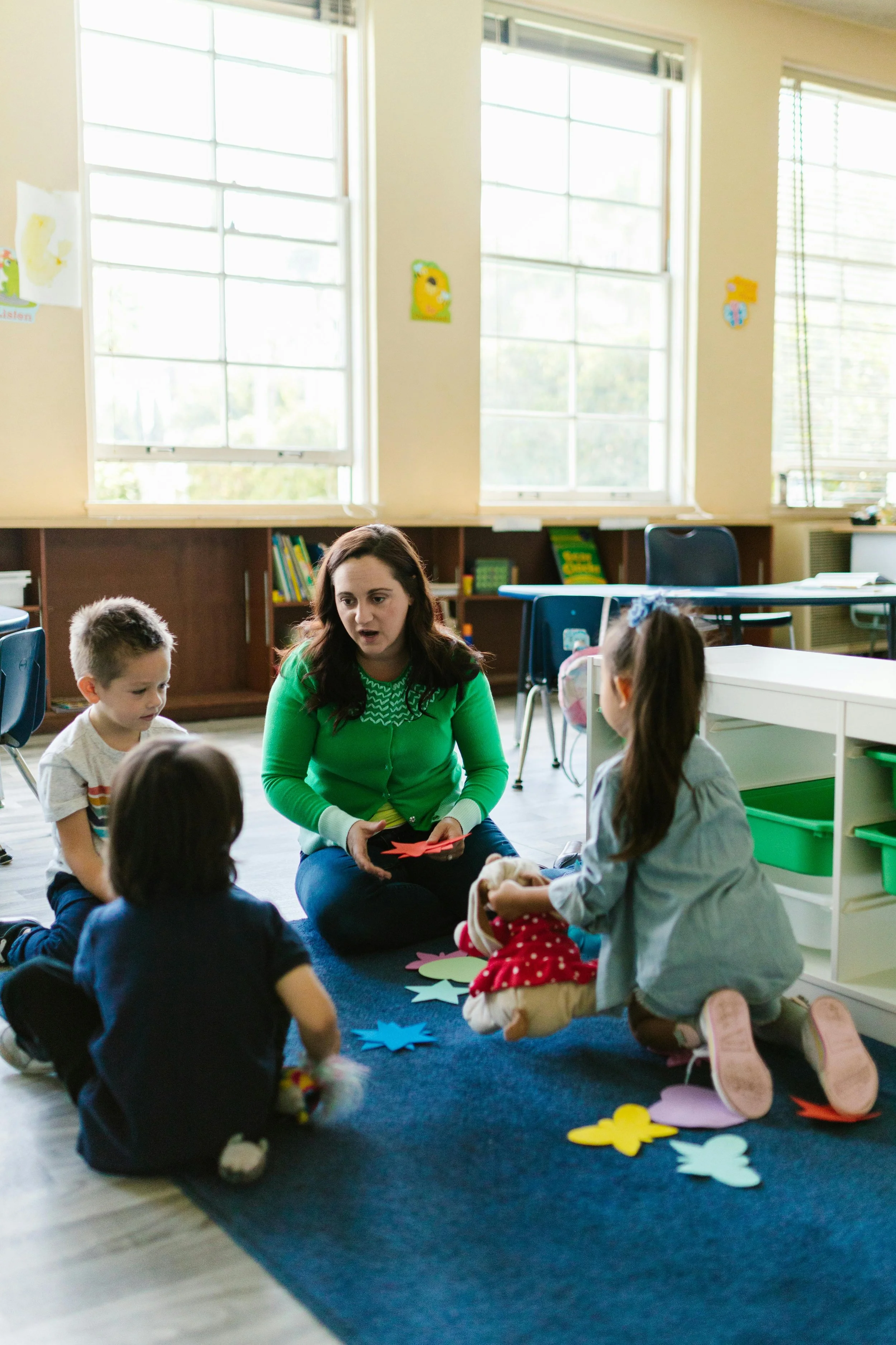 A teacher engaging with young children sitting on a blue carpet in a classroom, with colorful paper cutouts of flowers on the carpet.
