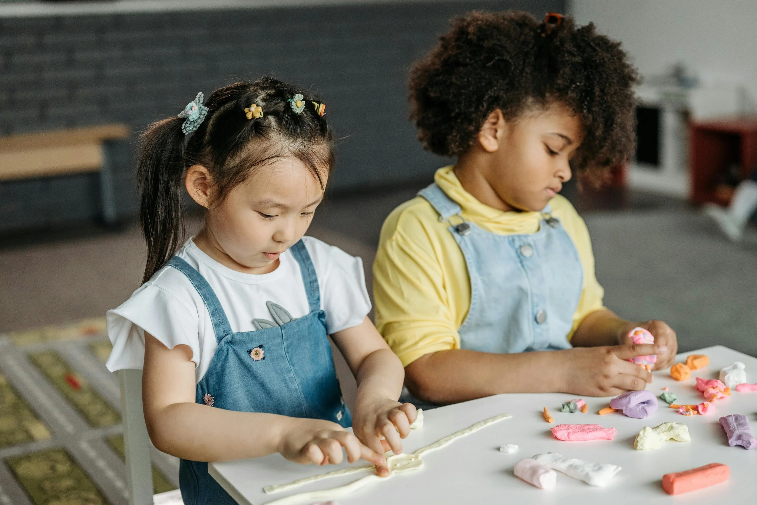 Two young children working with clay at a table, demonstrating hands-on sensory play, fine motor engagement, and emotional regulation in a classroom setting.