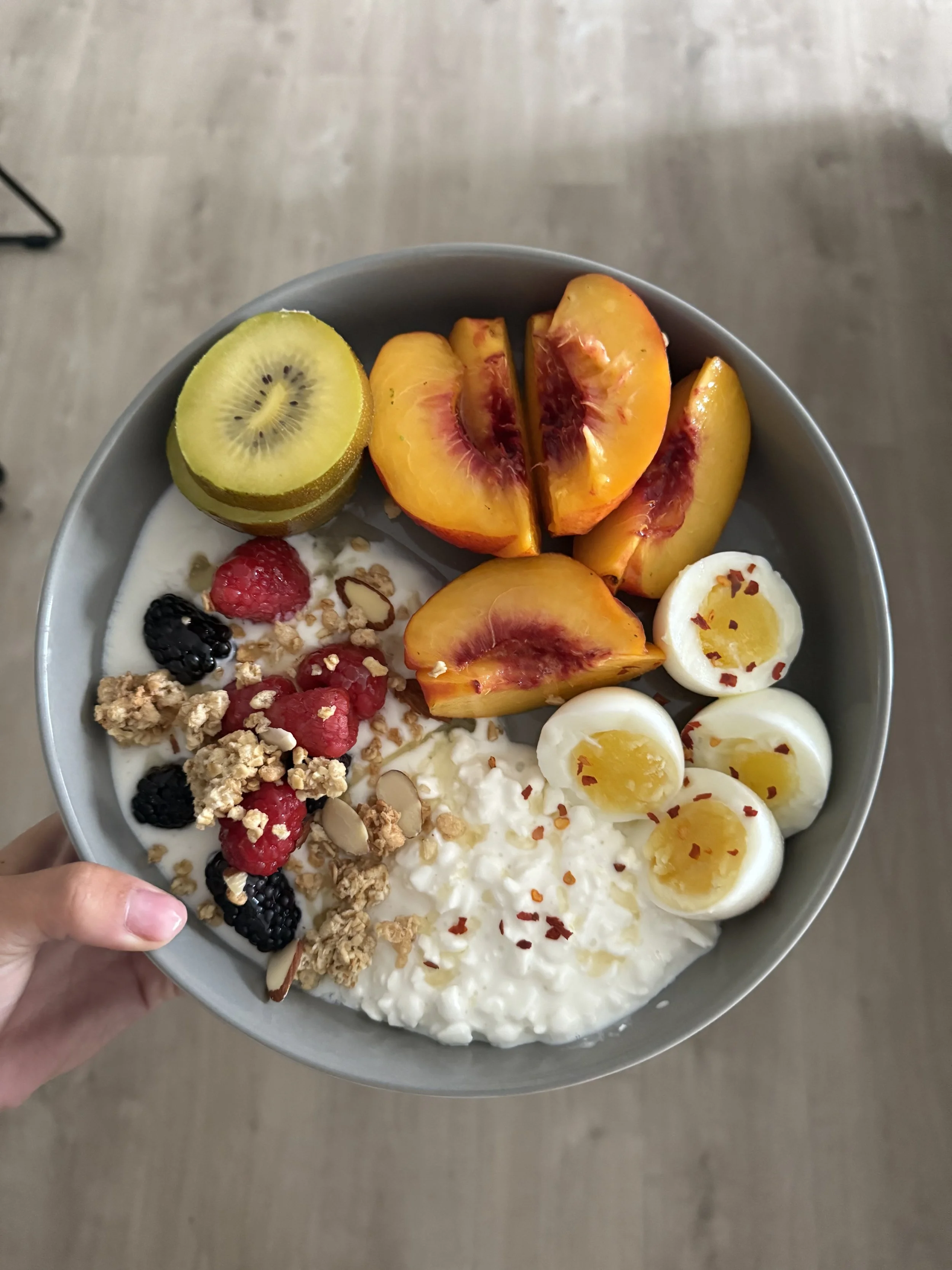 A bowl of yogurt topped with sliced kiwi, peaches, soft-boiled eggs, mixed berries, granola, and red pepper flakes, held in a person's hand over a wooden floor.