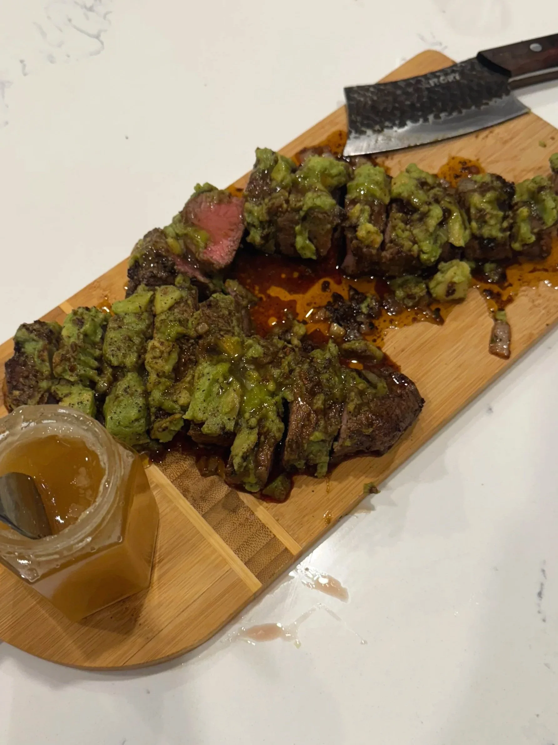 Sliced steak topped with green chimichurri sauce on a wooden serving board, with a jar of chimichurri sauce and a steak knife.