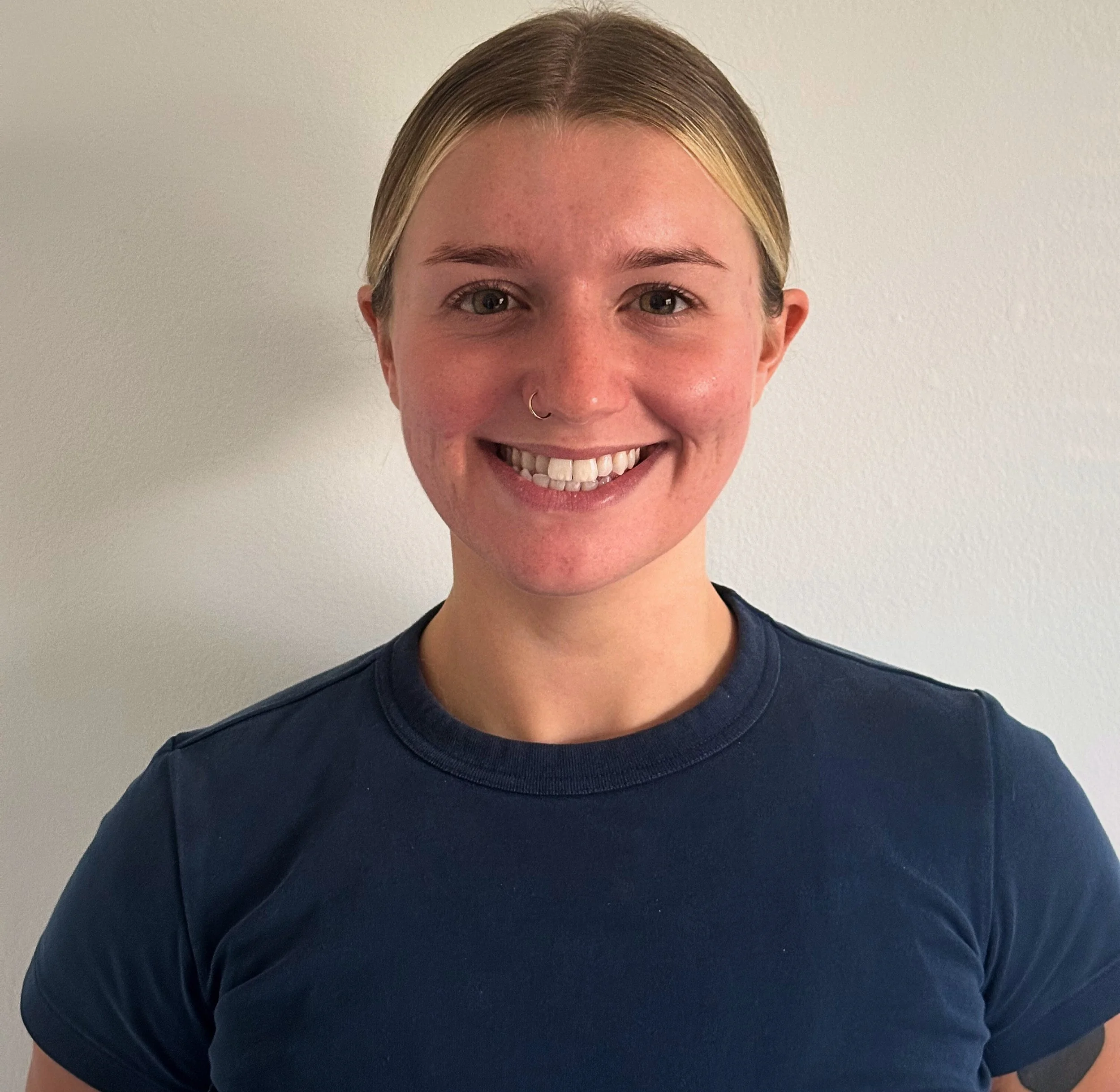 A young woman with blonde hair tied back, wearing a navy blue T-shirt, smiling against a plain white wall background.