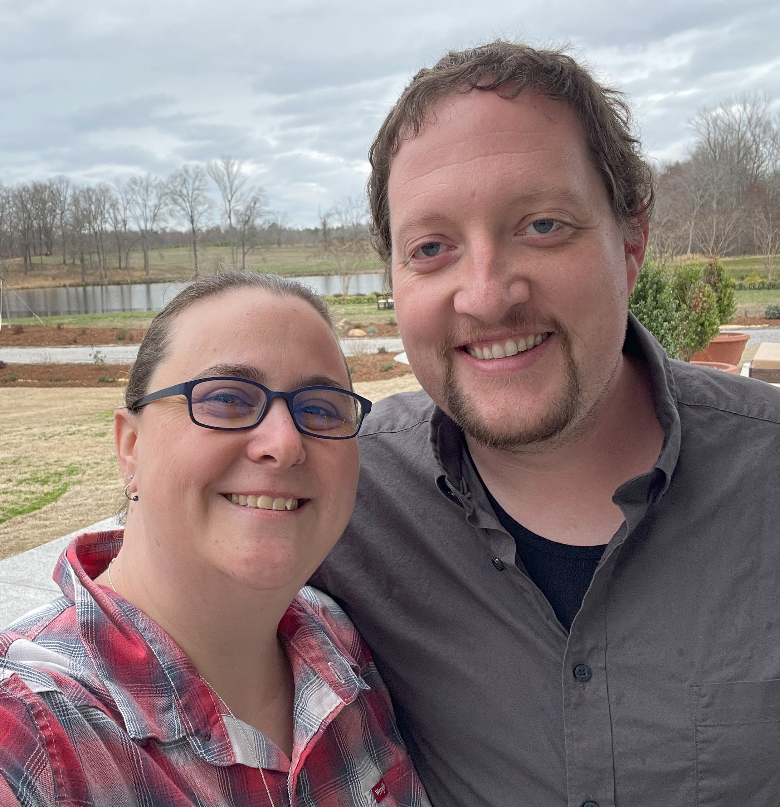 A smiling woman with glasses and a man with a beard taking a selfie outdoors with a pond, trees, and cloudy sky in the background.