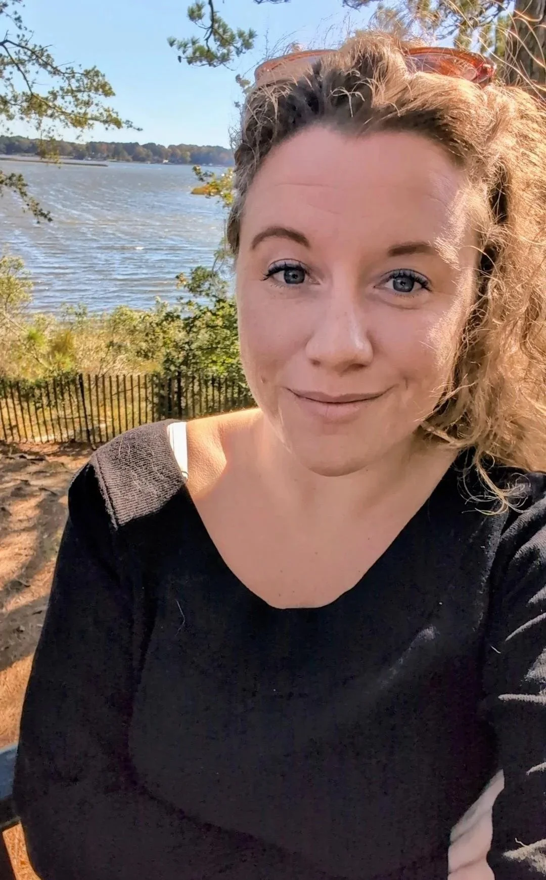 A woman with curly blonde hair taking a selfie outdoors near a body of water with trees in the background.