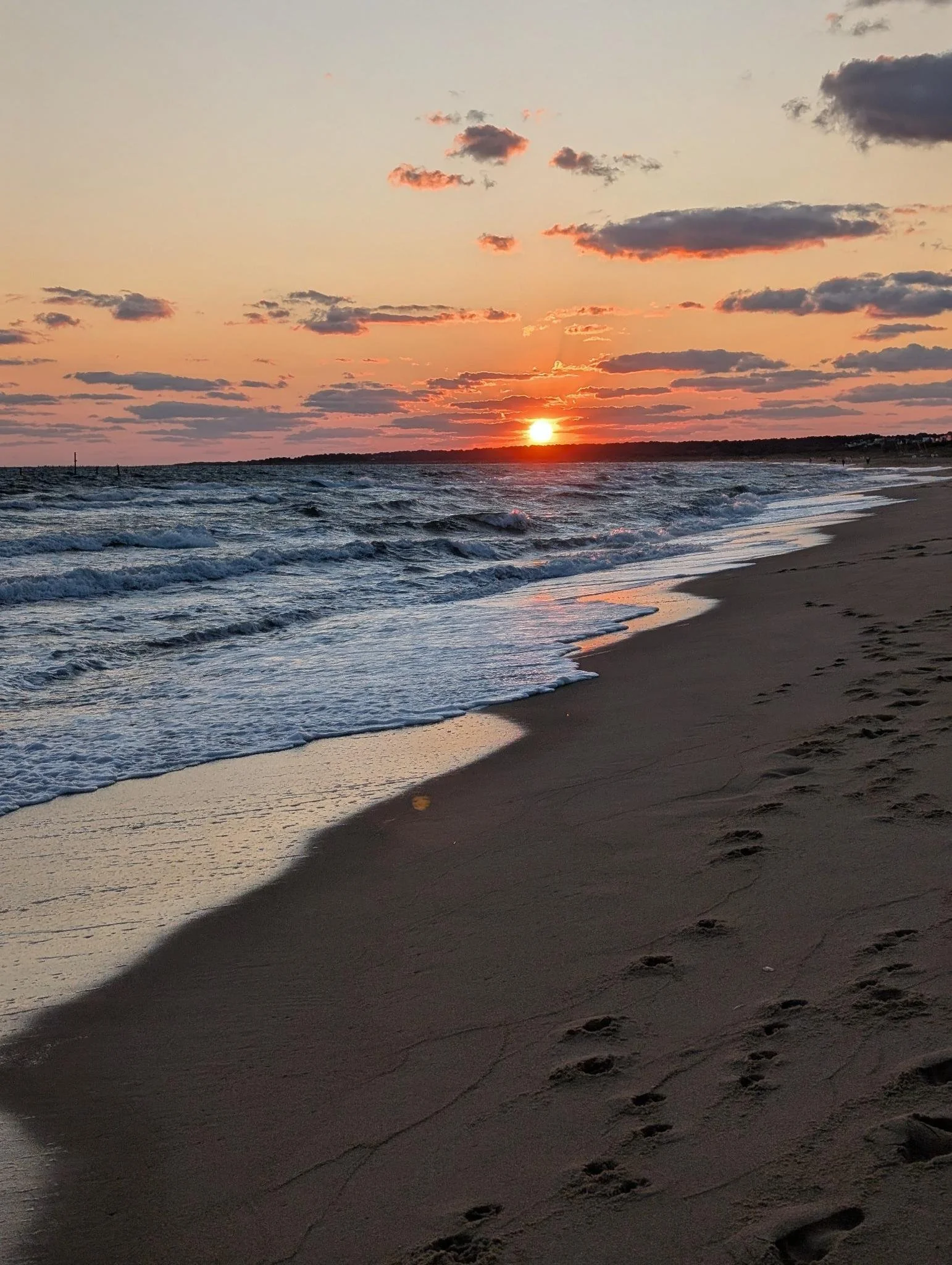 Sunset over the ocean with clouds, waves hitting the sandy beach, and footprints in the sand.