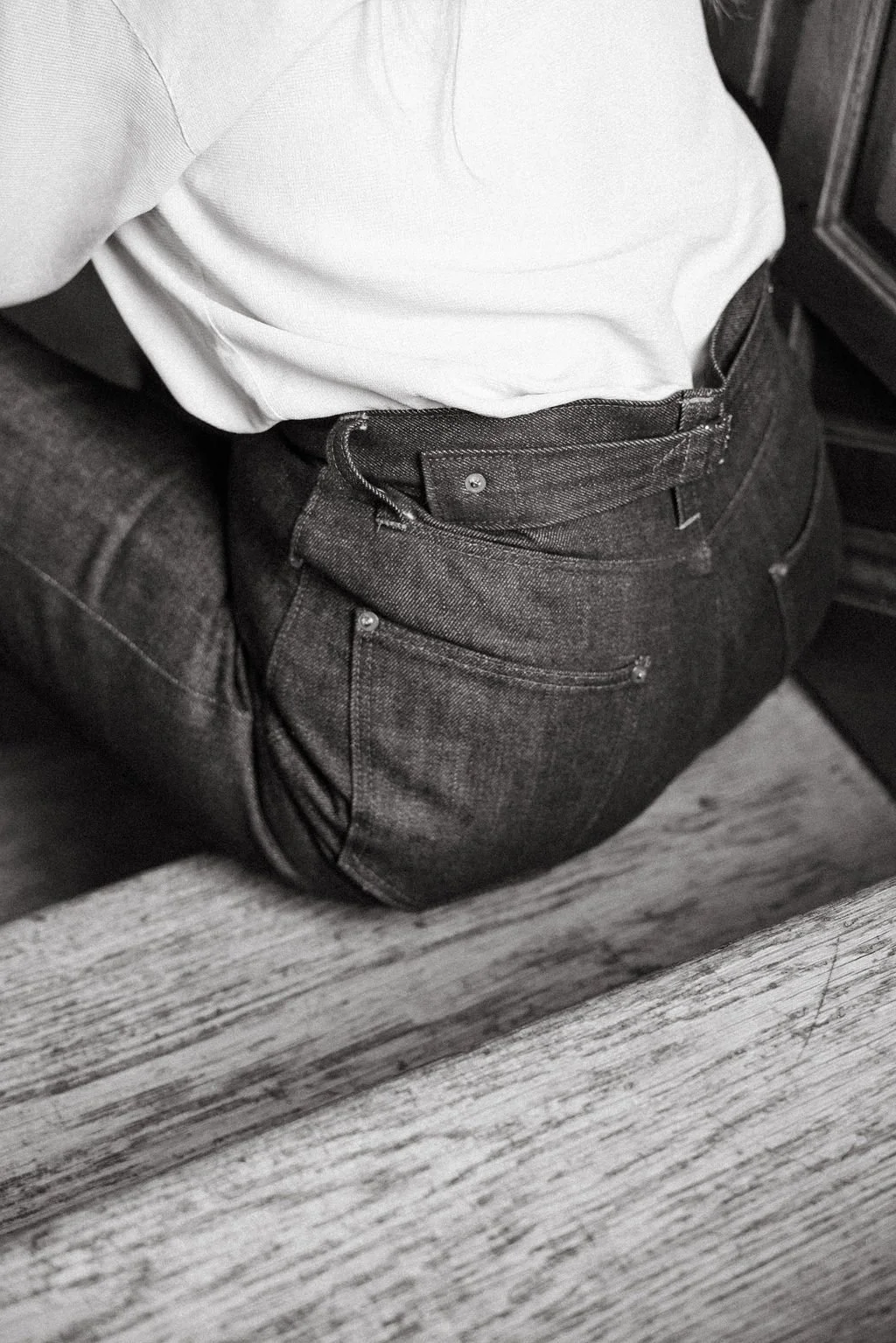 Close-up of a person wearing a white shirt and dark jeans, sitting on a dark-colored couch with a wooden floor underneath.