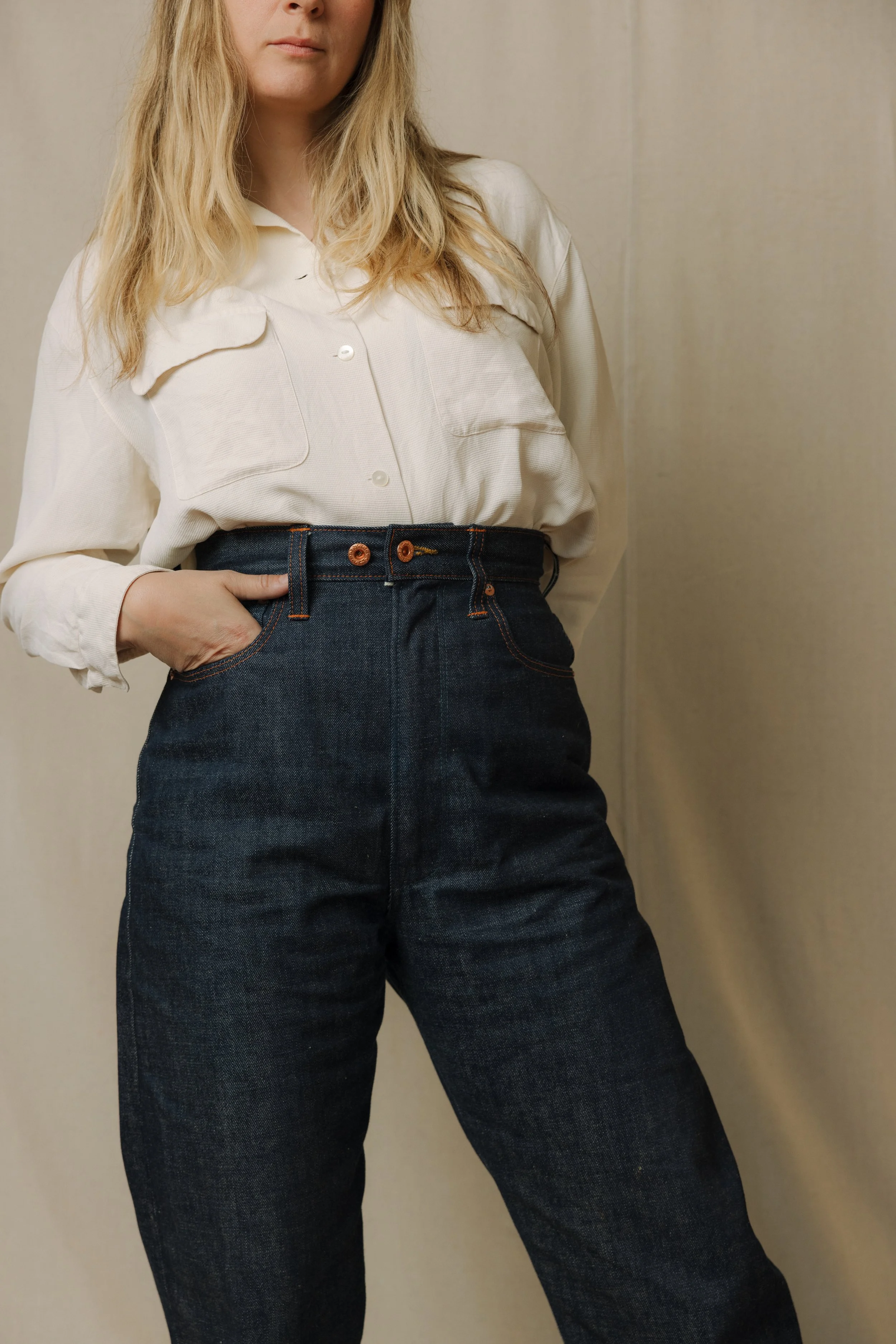 A woman wearing a cream-colored button-up shirt tucked into high-waisted dark denim jeans with orange stitching, standing against a neutral background.
