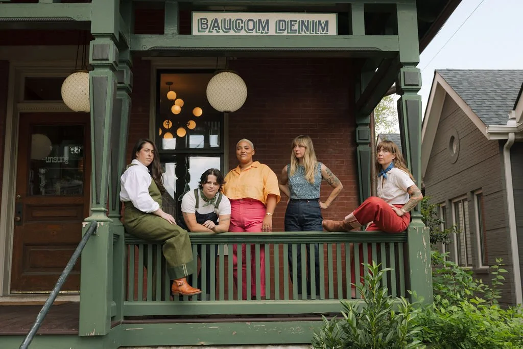 Five women sitting and standing on the porch of a building named 'Baucom Denim,' which has a green-painted, vintage-style wooden front with round paper lantern lights hanging inside.