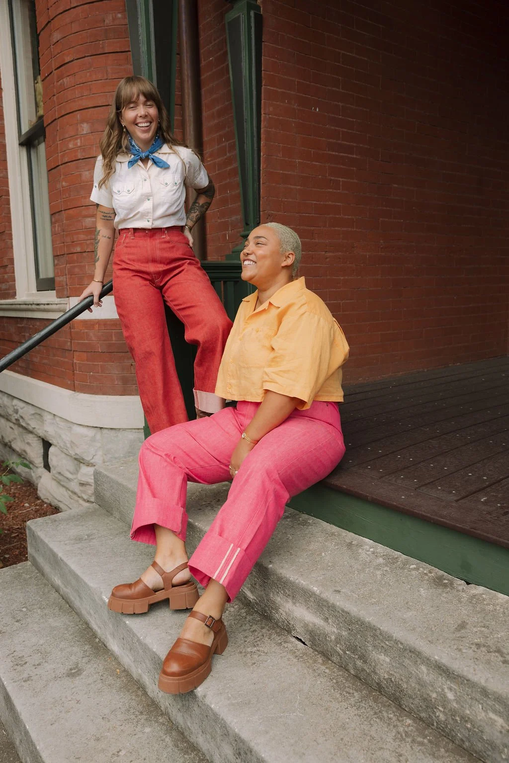 Two women smiling and chatting outside a brick building, one sitting on the stairs wearing a yellow top and pink pants, the other standing on the stairs wearing a white shirt, red pants, and a blue bandana.