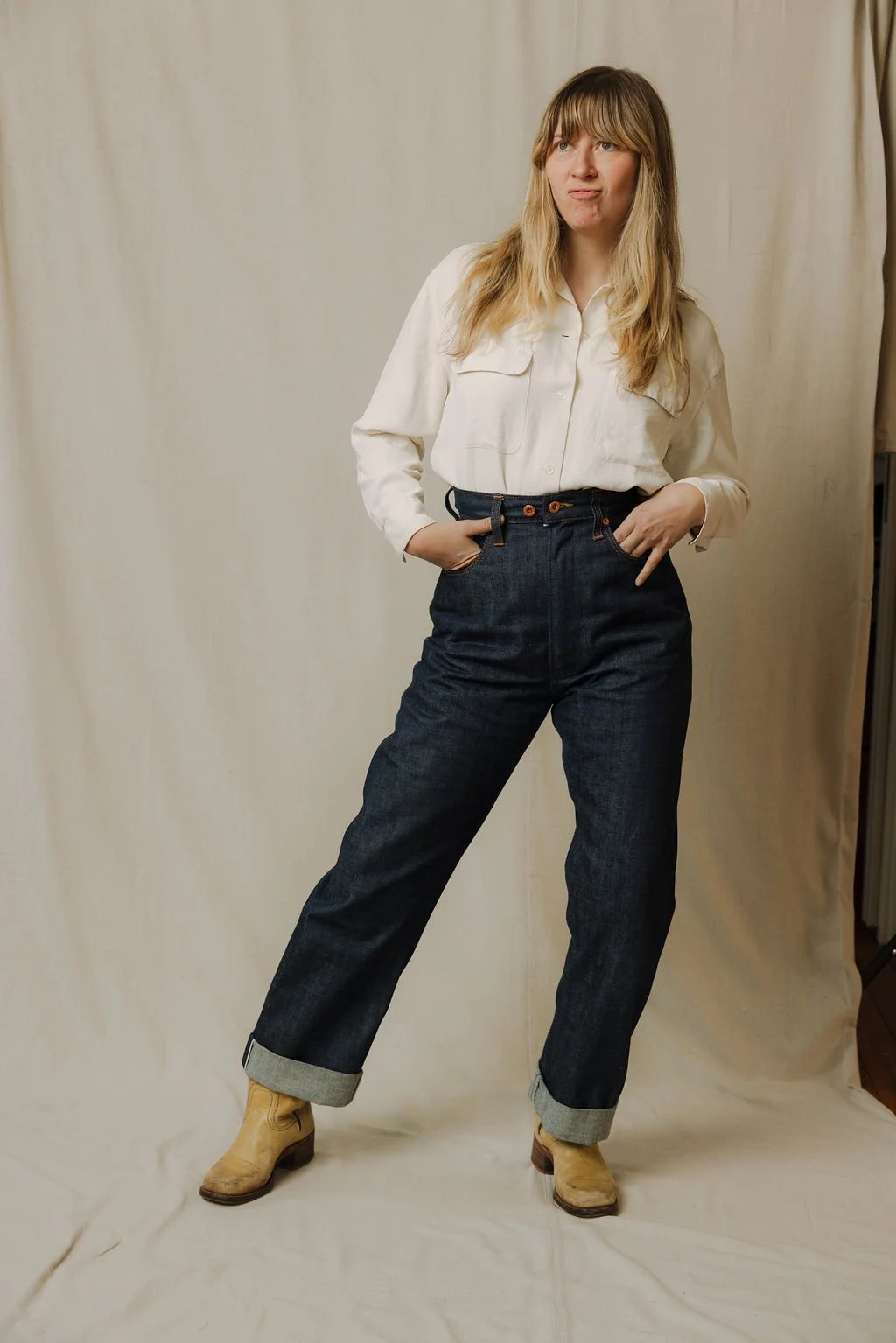 Woman with blonde hair wearing a white shirt, high-waisted dark jeans, and tan boots, standing with hands in pockets, in front of a plain beige backdrop.