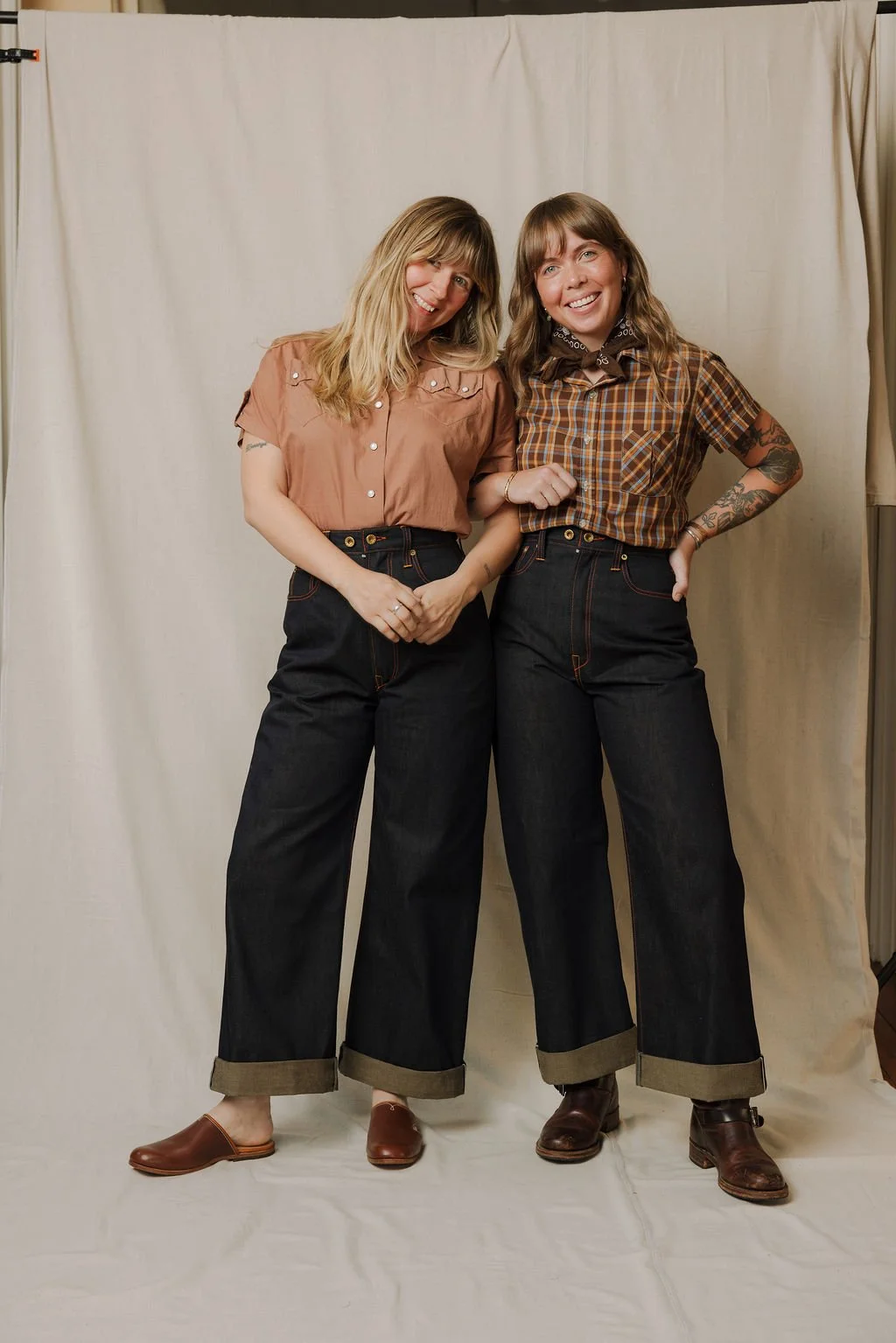 Two women standing close together, smiling, in front of a beige backdrop. They are wearing vintage-style outfits with high-waisted jeans and patterned shirts, one with a bandana and tattoos visible on her arm.