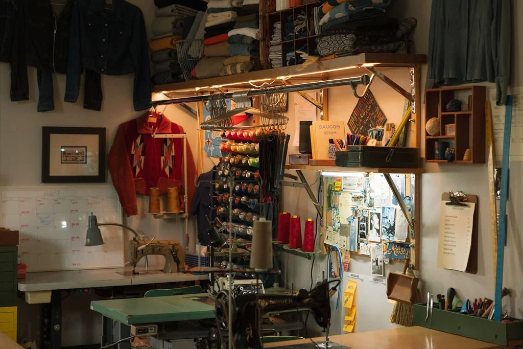 Sewing workspace with fabric, thread, and sewing machine, with shelves of folded fabric and clothing, and a whiteboard with notes.