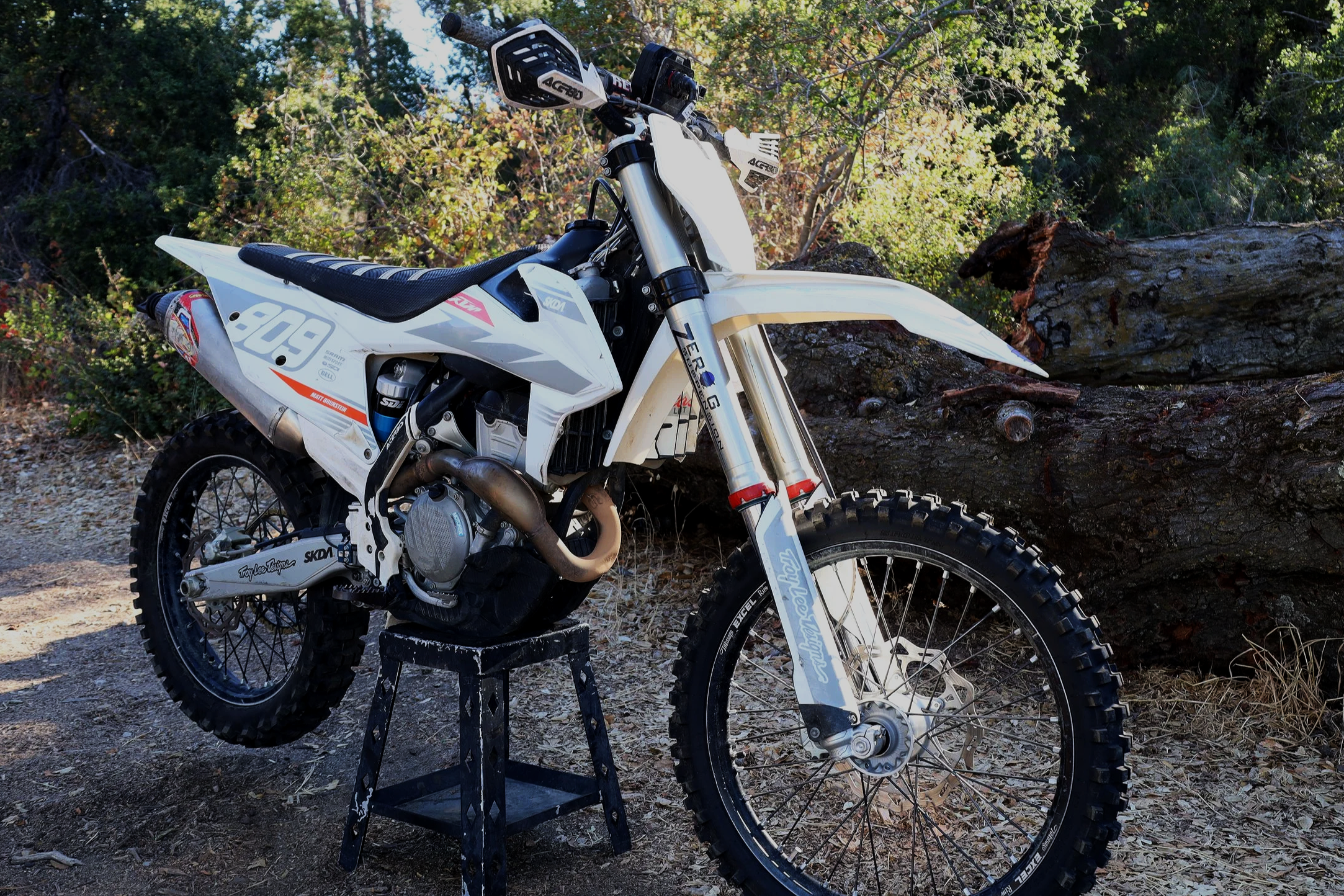A white off-road dirt bike resting on a stand outdoors on a dirt path, with trees and foliage in the background.
