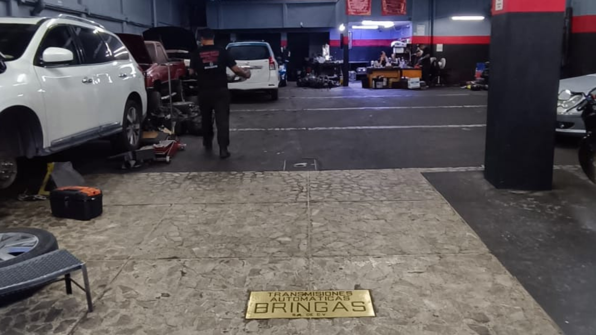 Interior de un taller mecánico con varios autos en reparación, herramientas y una persona trabajando con un vehículo.