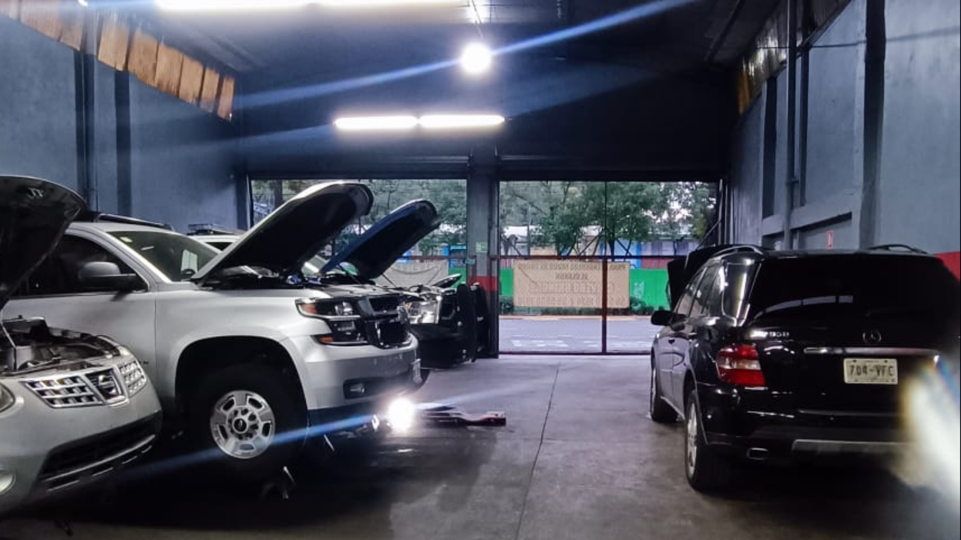 Autos estacionados en un taller, algunos con capós abiertos, en un espacio cerrado con iluminación artificial y vista a la calle a través de una puerta traslúcida.