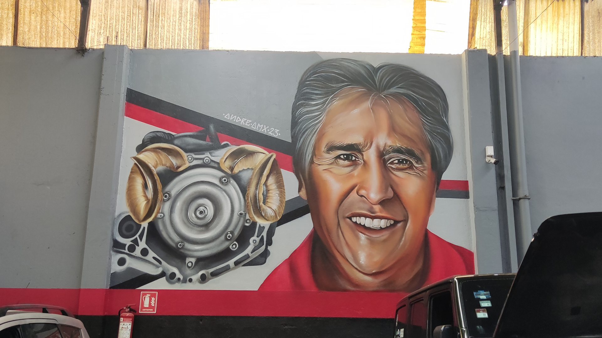 Mural que muestra la cara de un hombre sonriendo, con detalles de un motor de motocicleta y guantes dorados en el lado izquierdo, en un fondo gris con líneas rojas y negras.