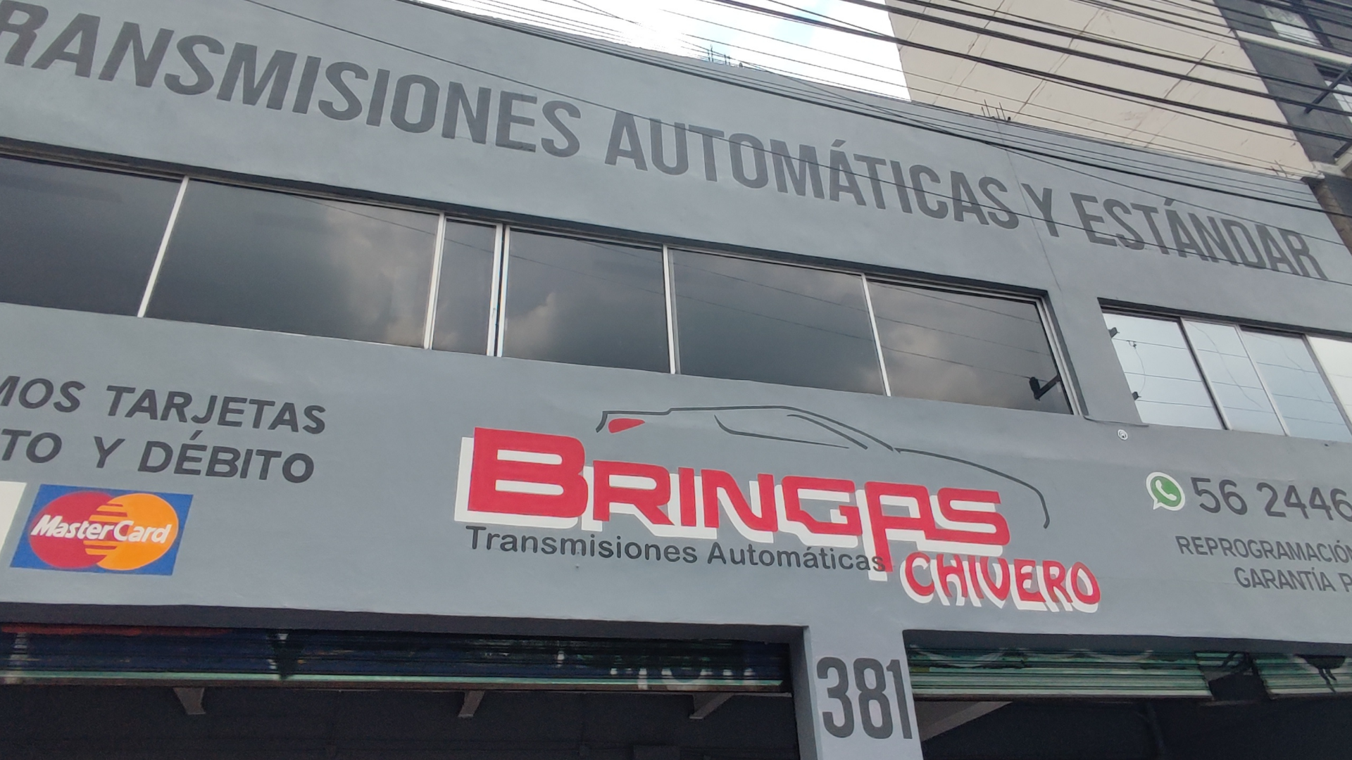 Fachada de una tienda de transmisiones automáticas llamada 'Bríngas Chívero', con logotipo, anuncios de servicios y pago con tarjetas Mastercard y opciones de contacto vía WhatsApp.
