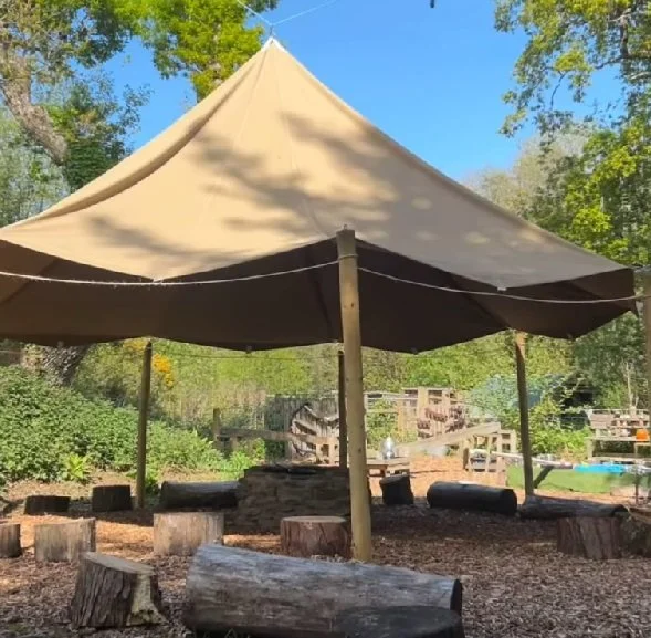 A beige outdoor canopy supported by wooden poles, with tree stump seats arranged around a stone firepit in a wooded area.