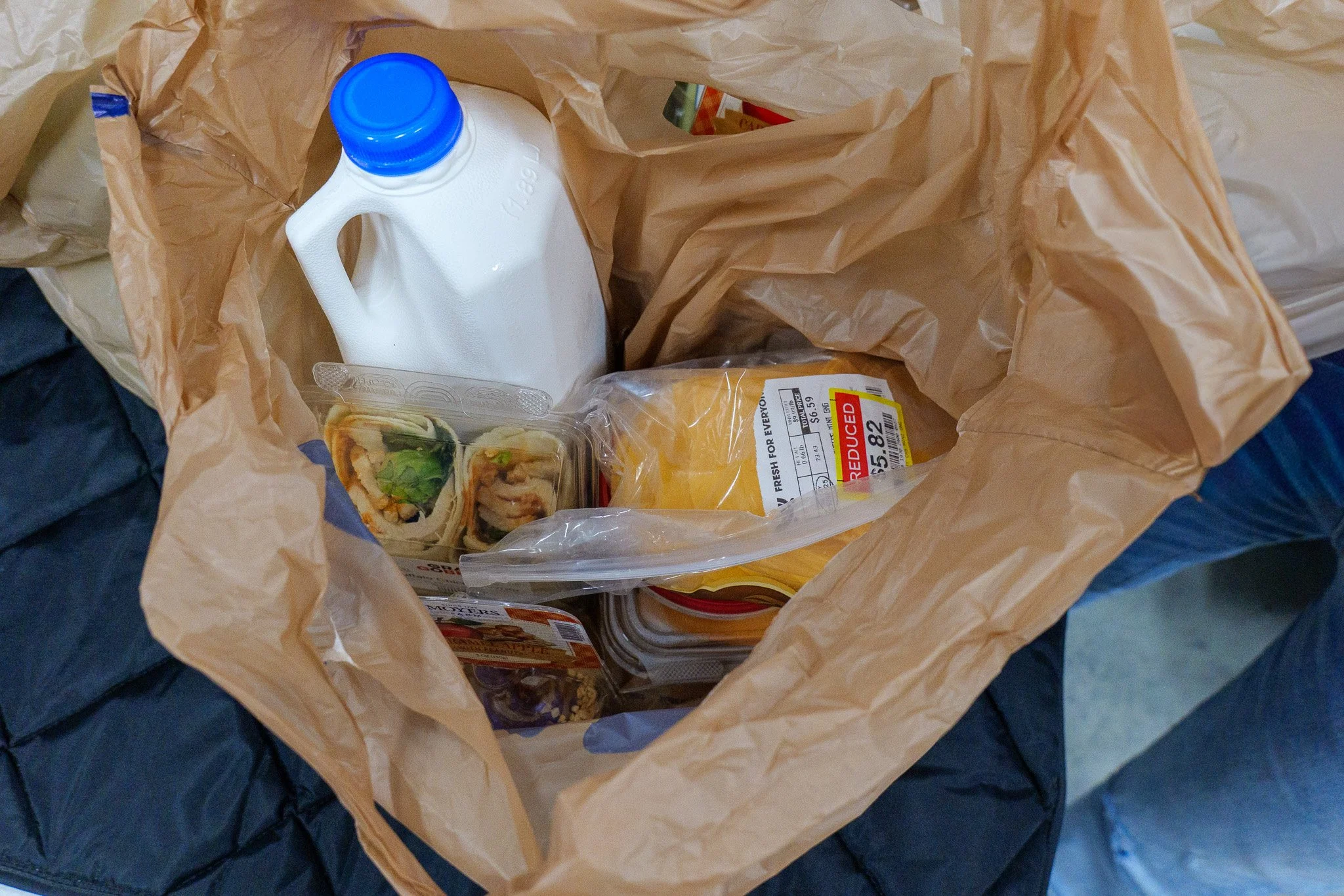 Inside a brown paper grocery bag, there is a gallon of milk, a package of sliced bread, and several food containers including sushi and pre-packaged meals.