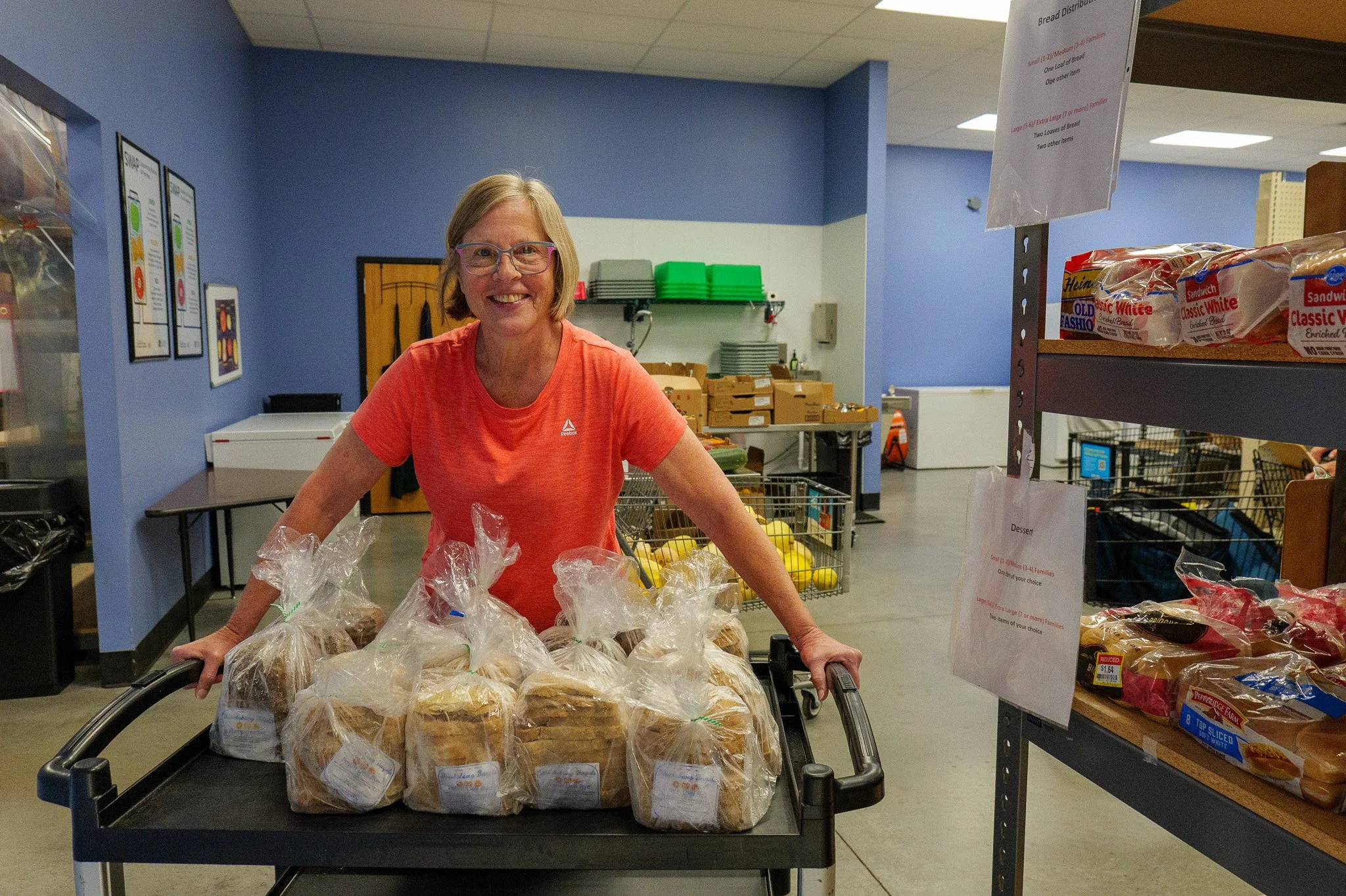 Woman smiling at the camera with a cart full of pre-packaged bread in a food pantry or grocery store setting.