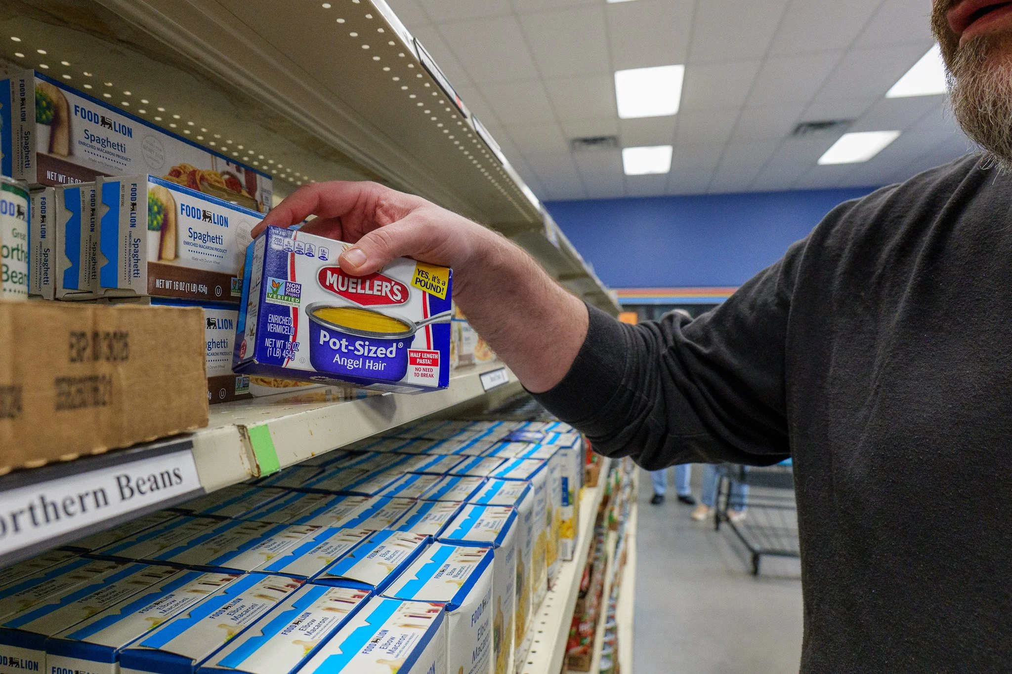 Person selecting a can of Mueller's pot-sized angel hair pasta from store shelf in aisle with canned goods.