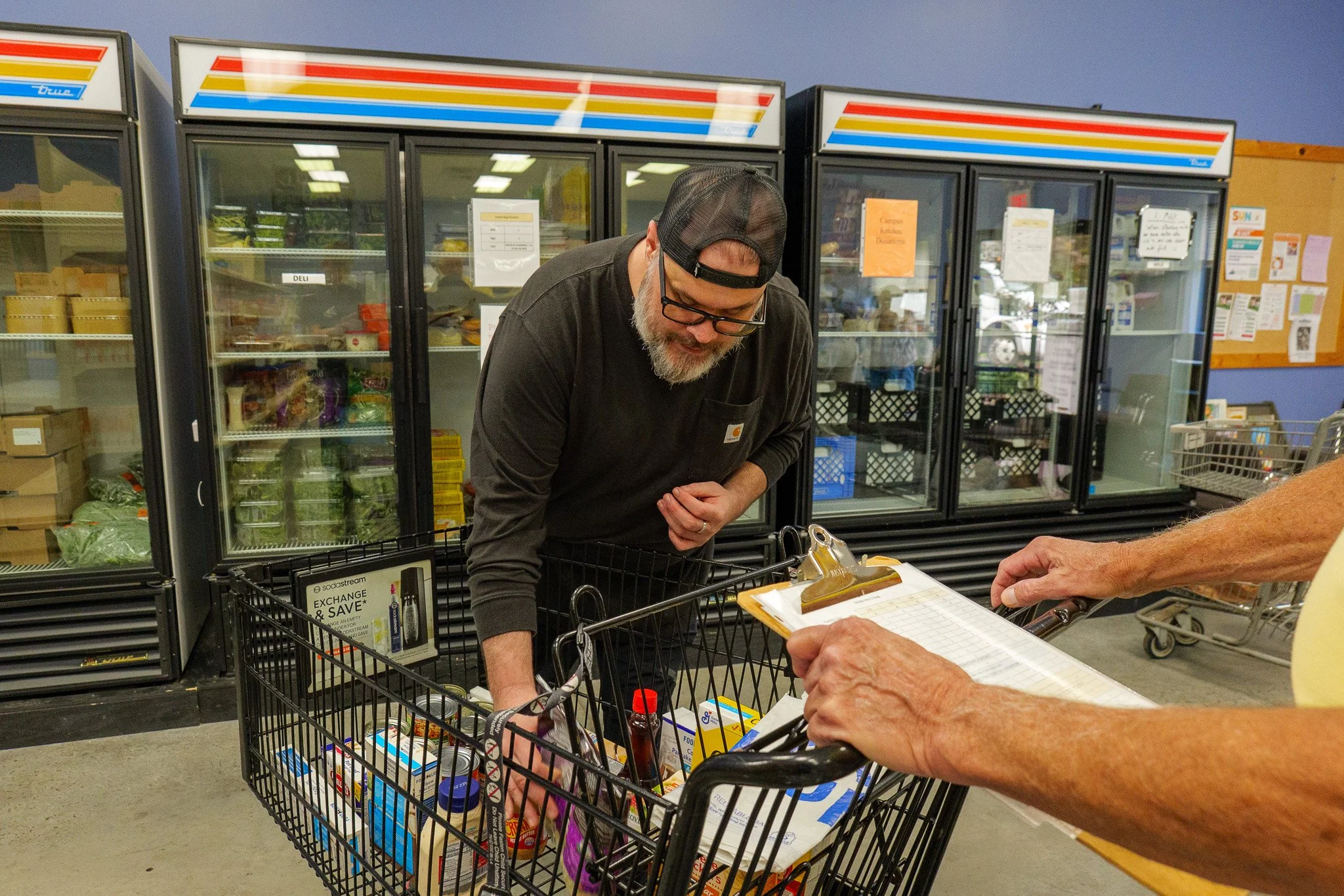 A man with glasses, a beard, and a black cap shopping in a grocery store, looking into the shopping cart with products. Another person is holding a clipboard or shopping list near the cart.
