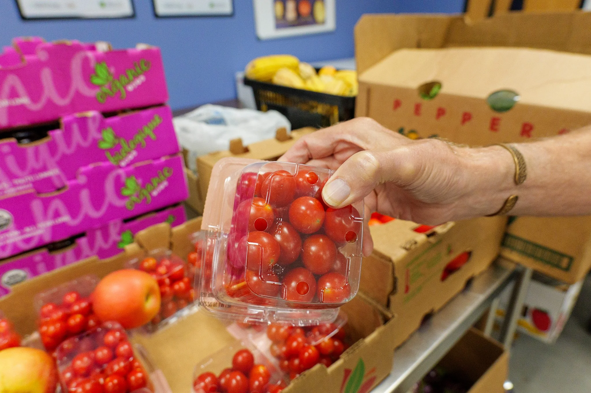 A person's hand holding a plastic container of cherry tomatoes in a grocery store produce section. There are various fruits and produce boxes on display.
