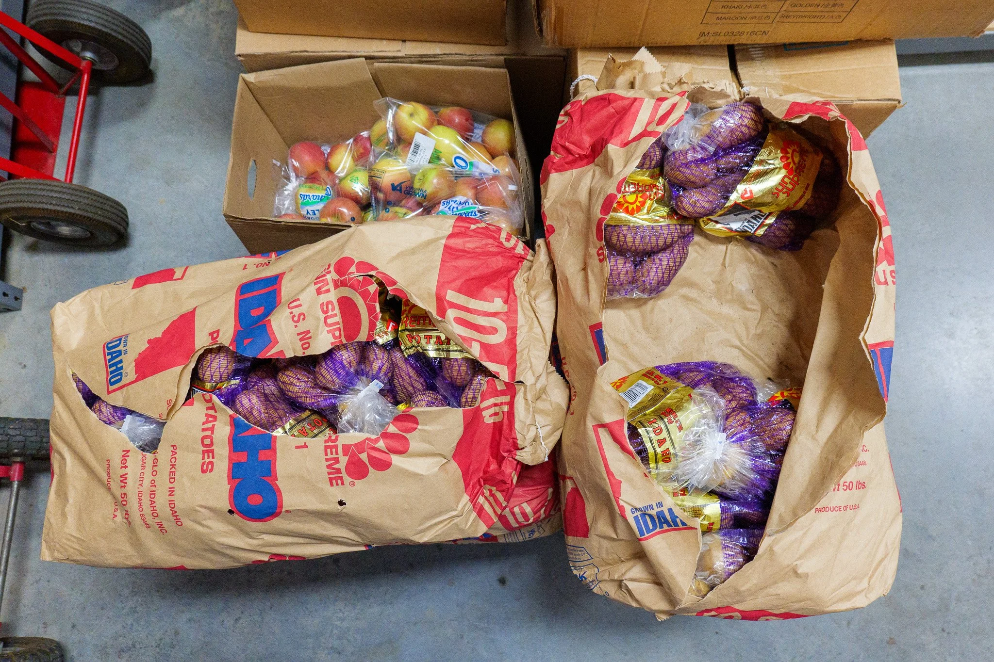 Three large brown paper bags filled with Idaho potatoes, two bags contain purple potatoes, and a cardboard box of apples, on a concrete floor.