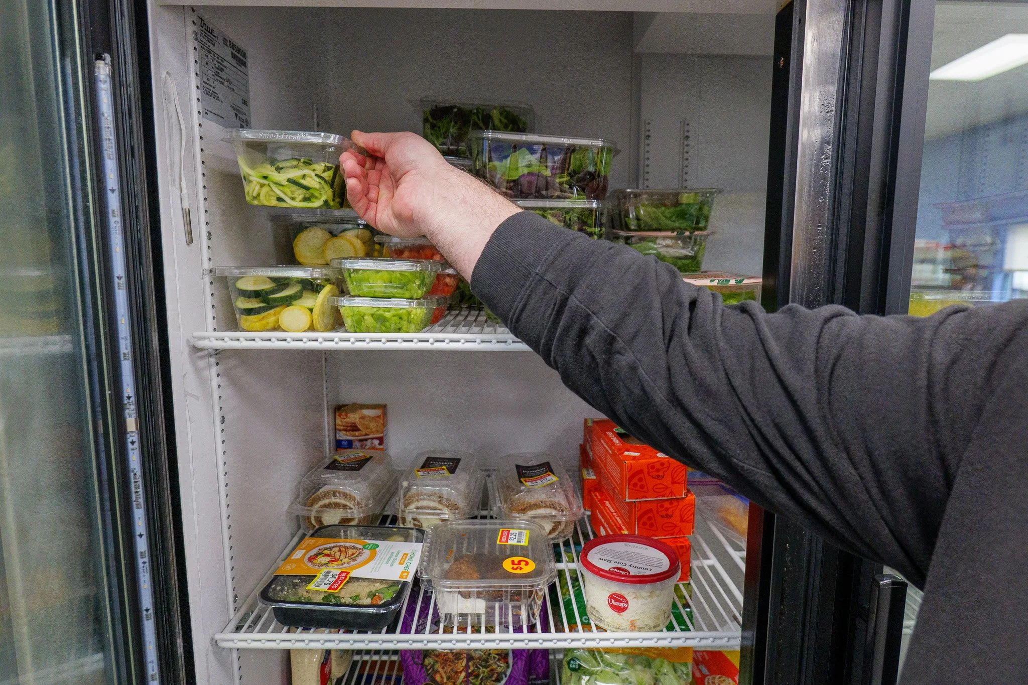 Person's arm reaching into a refrigerator, placing a plastic container of zucchini noodles on a shelf filled with various pre-packaged salads and food items.