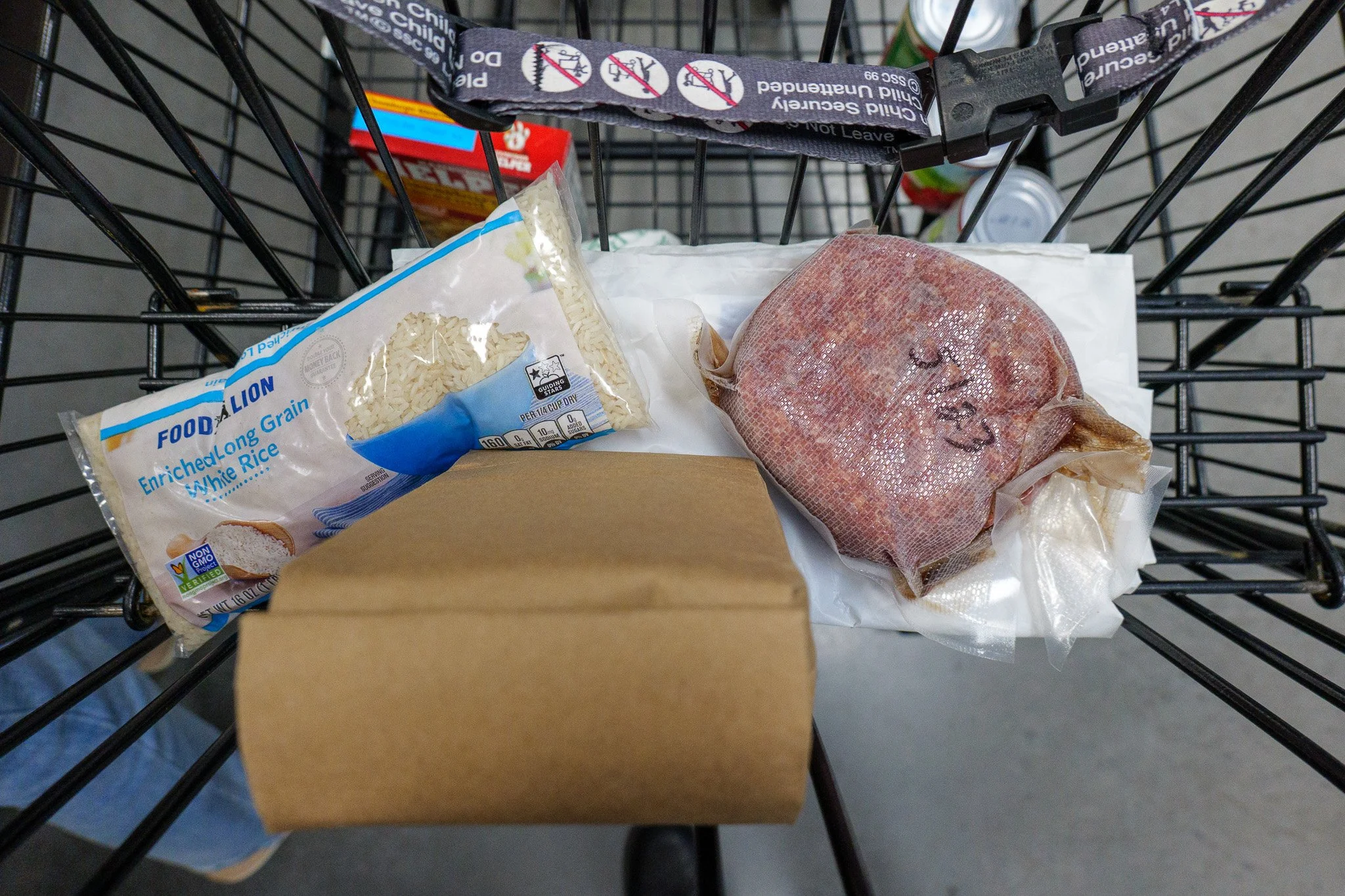 A grocery cart containing a package of white rice, a wrapped piece of meat in plastic, and other food items visible in the background.