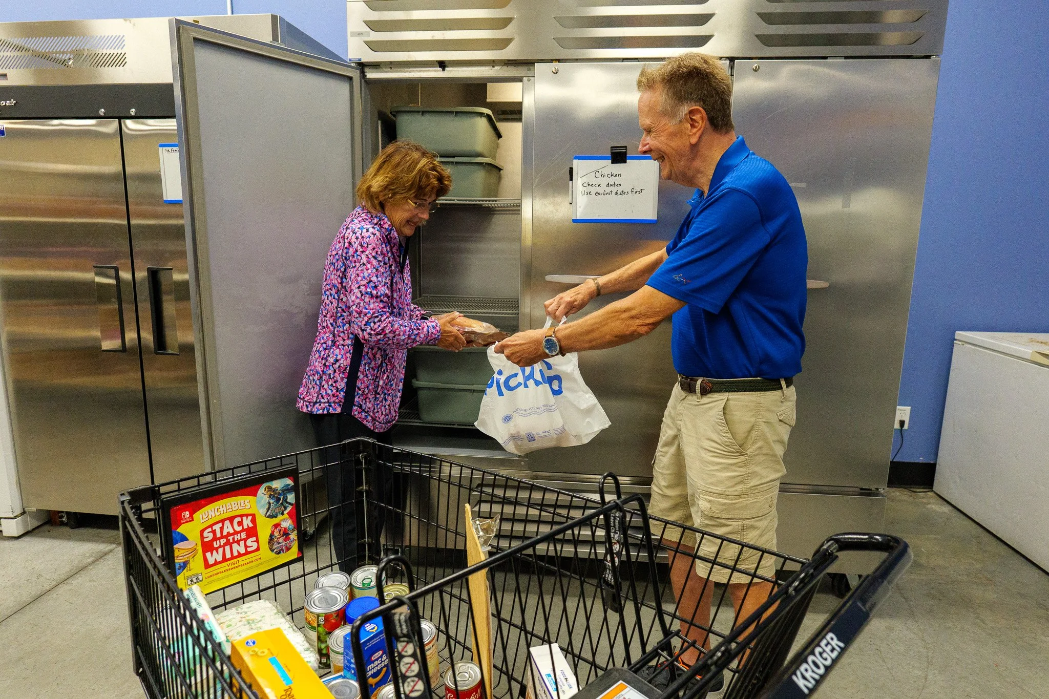 A man in a blue polo shirt and tan shorts is handing a bag of groceries to a woman with glasses and a colorful patterned jacket in a grocery store aisle, with a shopping cart in the foreground and a stainless steel fridge behind them.