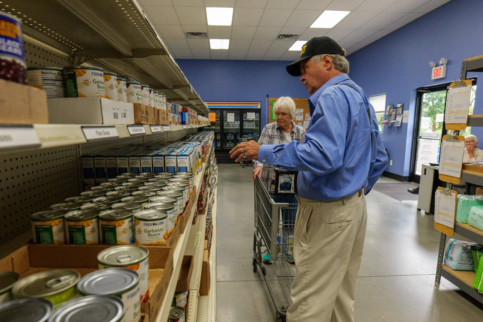 An elderly man and woman shopping in a grocery store aisle with canned food and soup on shelves, man pushing a shopping cart, store interior with fluorescent lighting and blue walls.