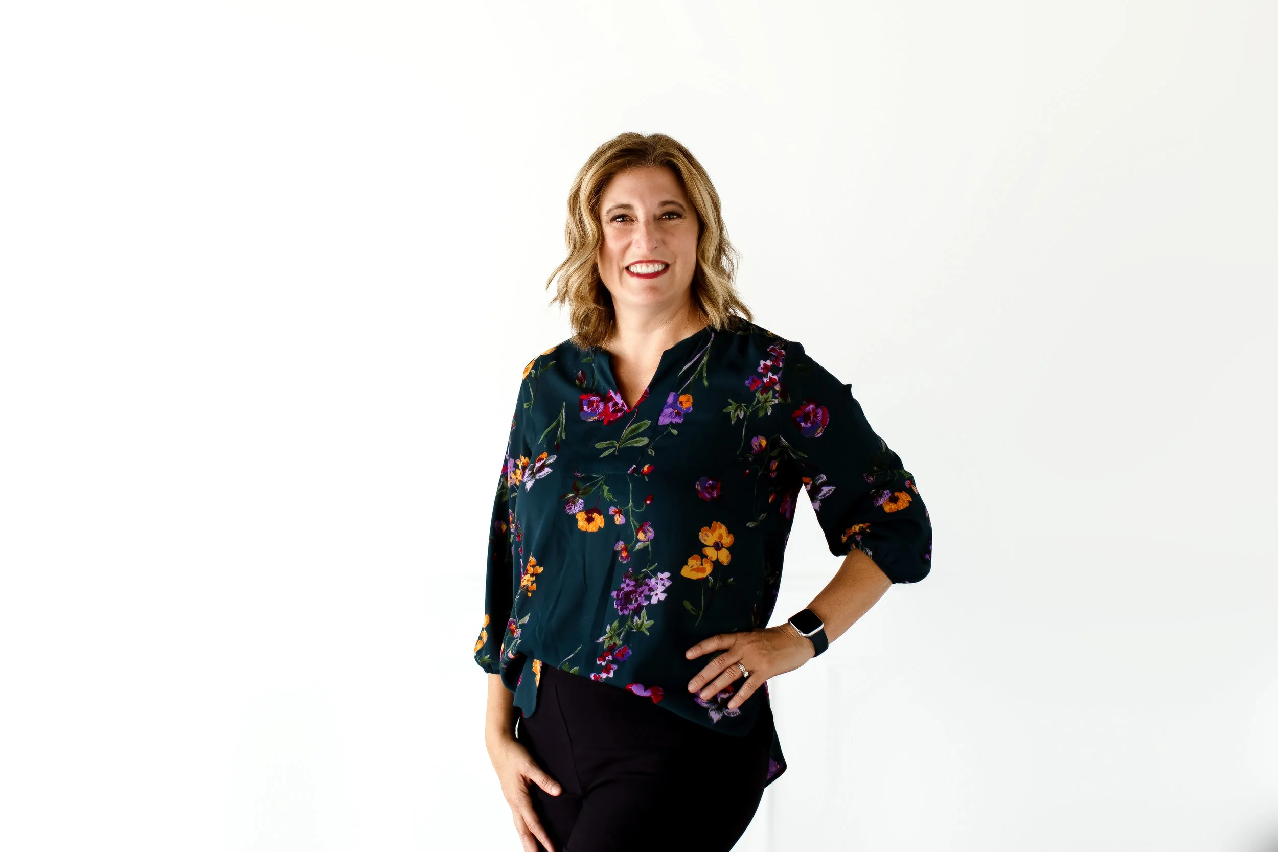 A woman with shoulder-length blonde hair, wearing a dark floral blouse, black pants, and a smartwatch, standing against a plain white background with a happy expression.
