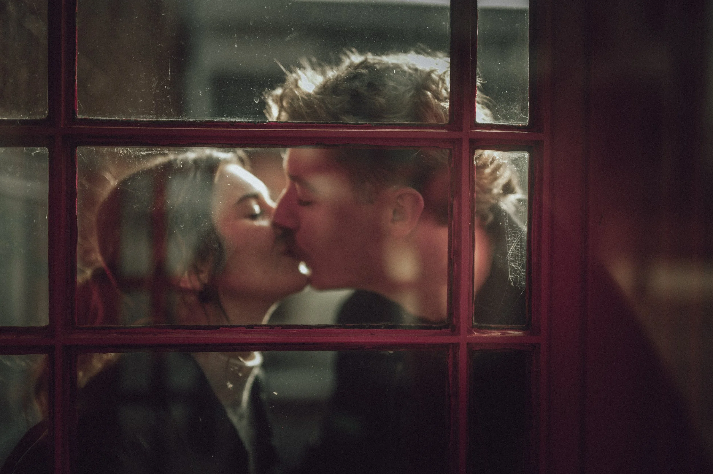 Engaged couple kissing through a glass door pane reflection maroon door