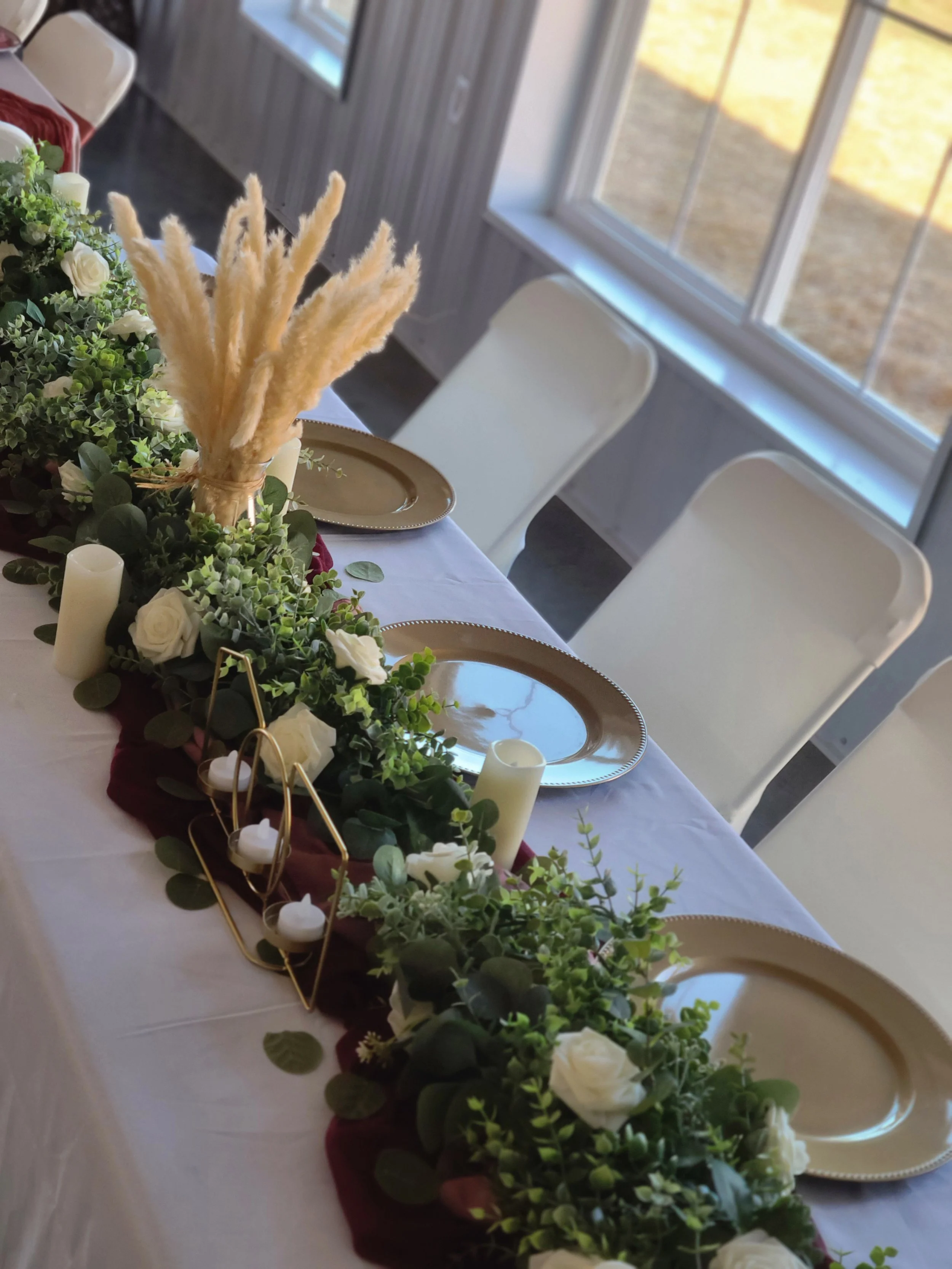Wedding table decor greenery runners gold candle holders flameless candles burgundy silk runners white foam flowers white table cloth white chair covers gold chargers pampas grass
