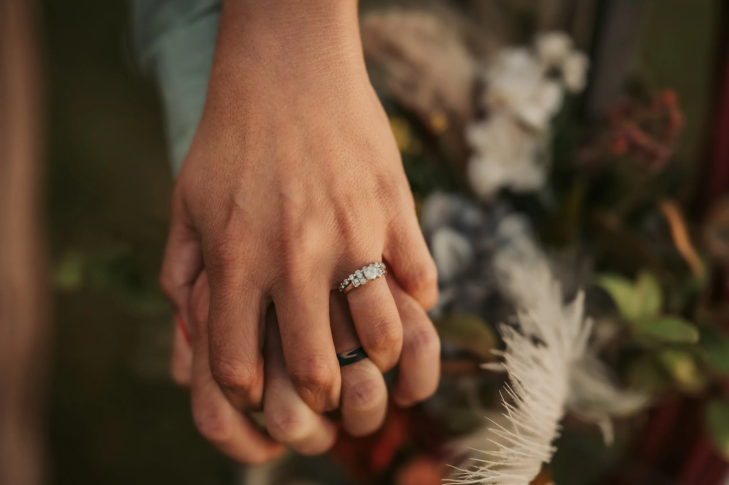 Couple holding hands engagement ring with band floral and feather background