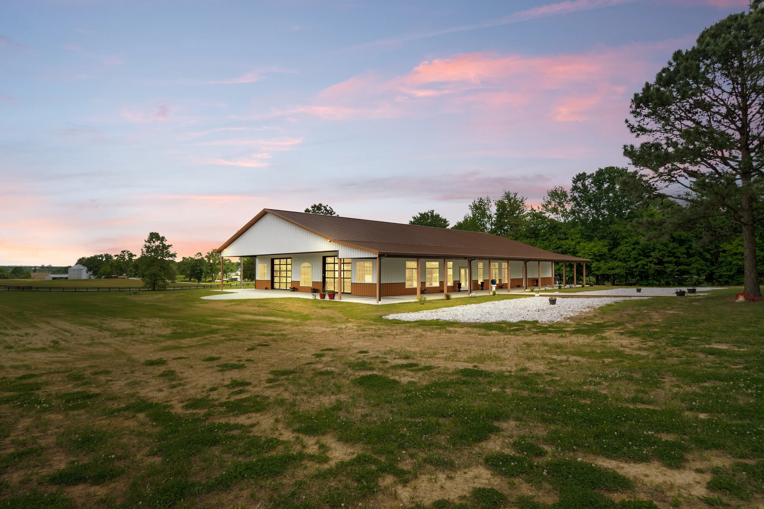Large white elegant building with copper roof 360 panoramic view from within the venue itself large glass doors mature trees green grass rock landscaping fencing around property flowers in pots blue and pink sky