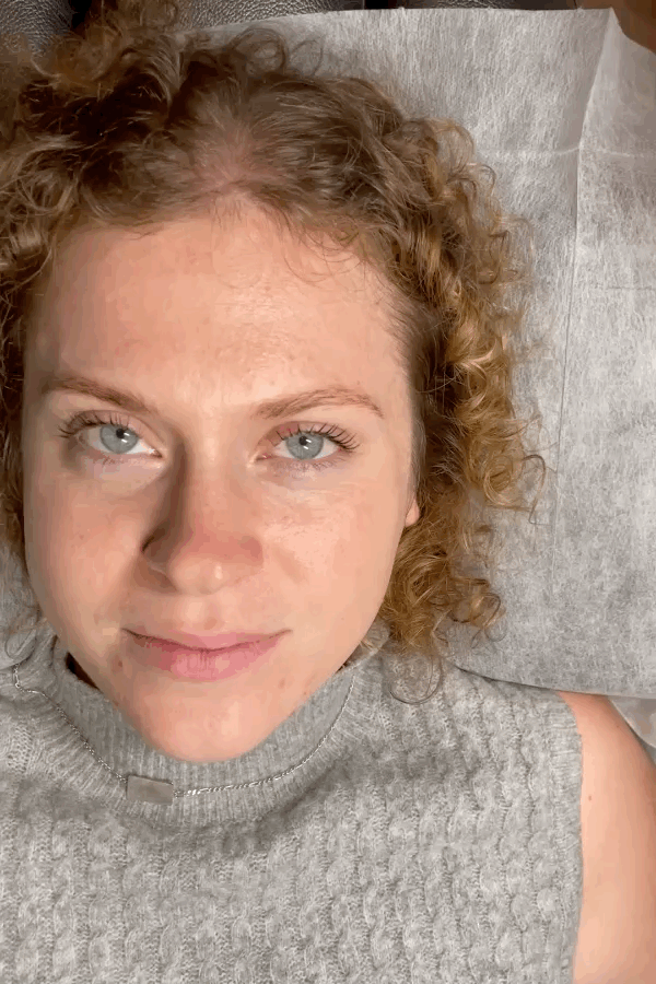Close-up of a woman with curly red hair and blue eyes lying on a bed, looking at the camera.