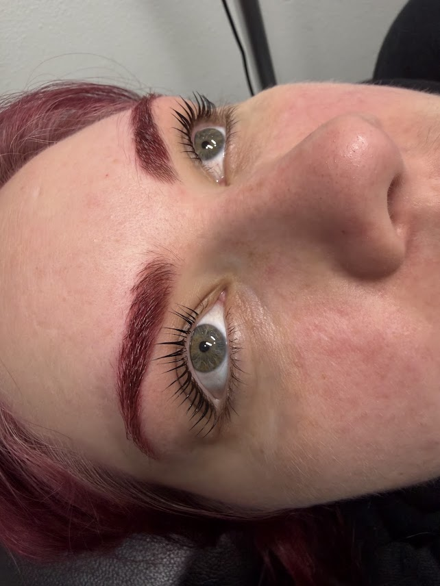 Close-up of a woman's face showing her eyes, with freshly painted red eyebrows and long, dark eyelashes, lying on a surface.