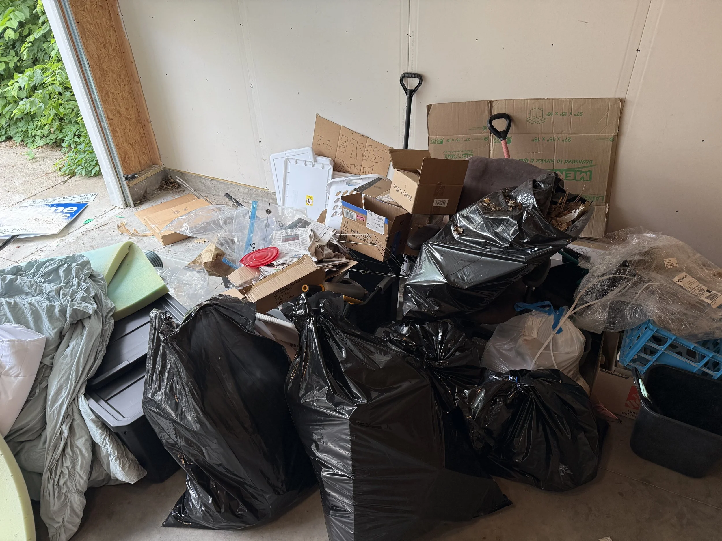 Cluttered garage with garbage bags, cardboard boxes, and miscellaneous items stacked on a table and on the floor.