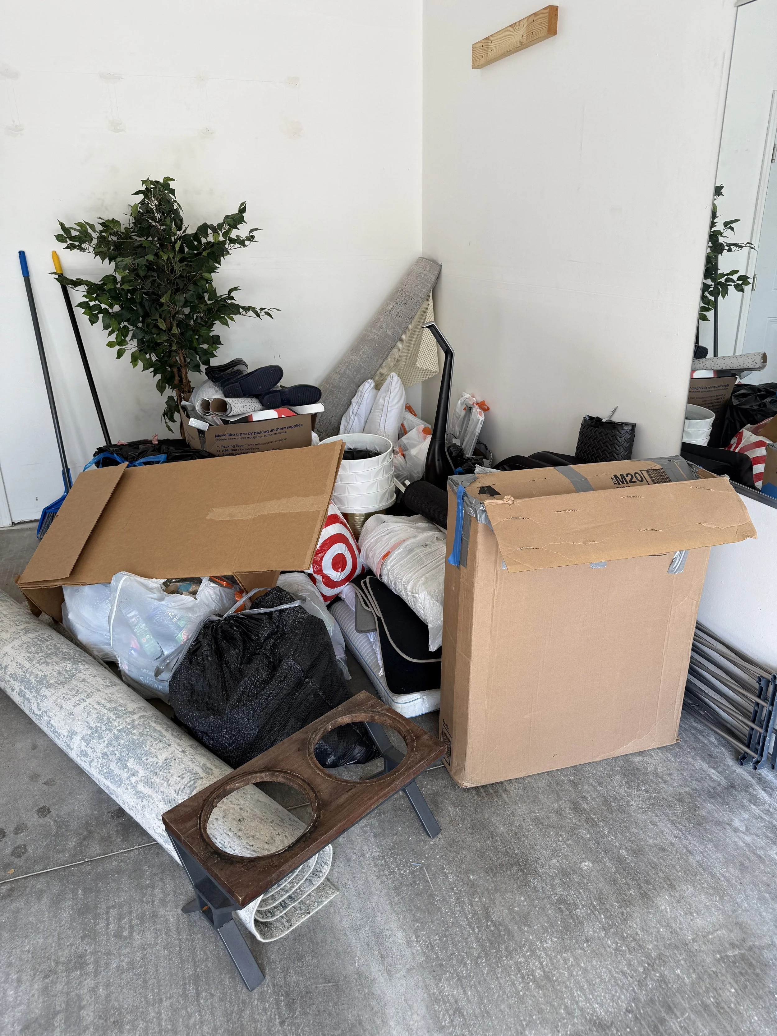 A cluttered garage corner with boxes, a rolled-up rug, a small table, pillows, and cleaning tools.
